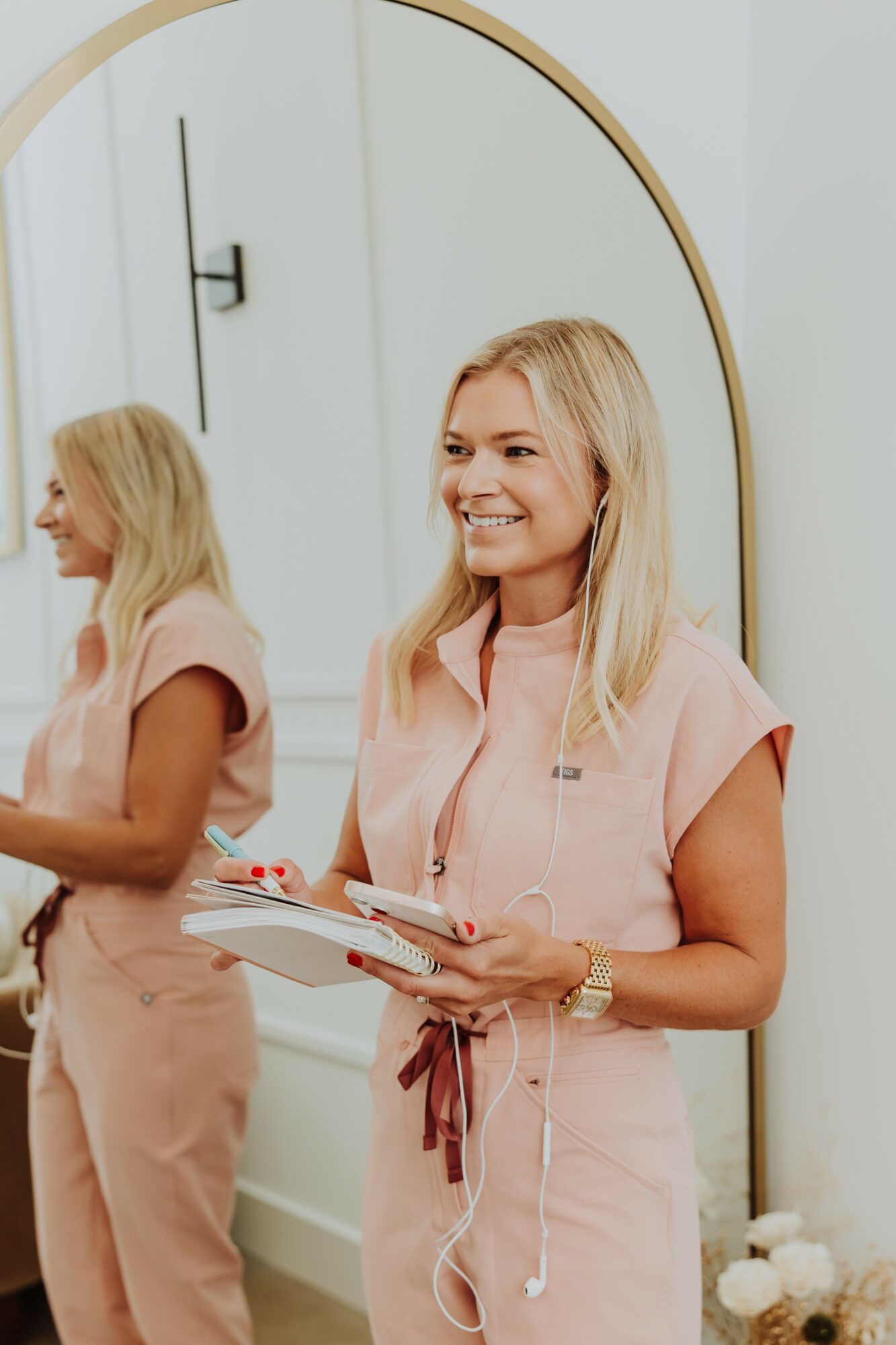 Woman with blonde hair smiling, holding a notebook, reflected in a mirror, wearing a pink outfit, in a bright room.