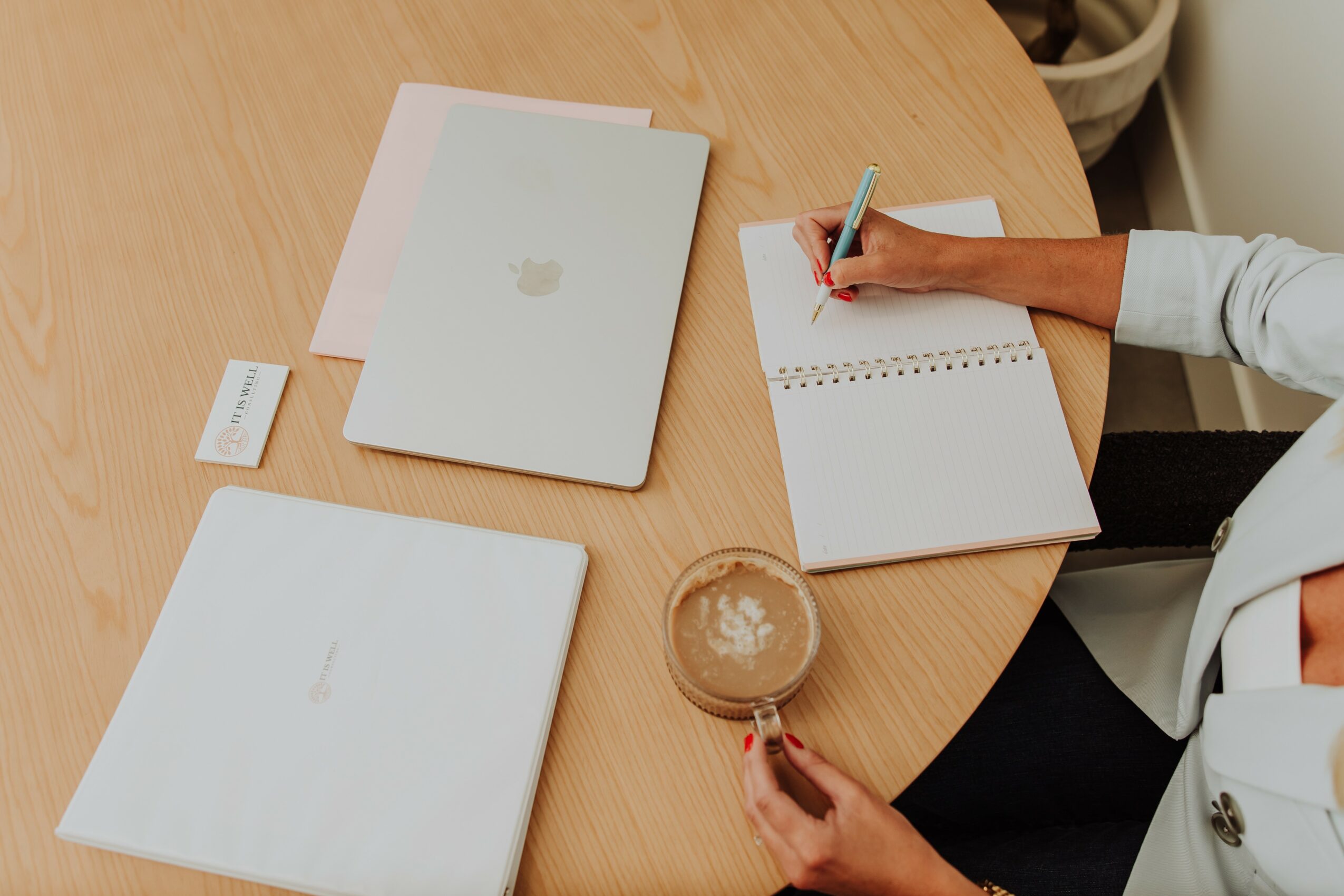 Person writing in a notebook on a wooden table with laptops and a glass of coffee nearby.