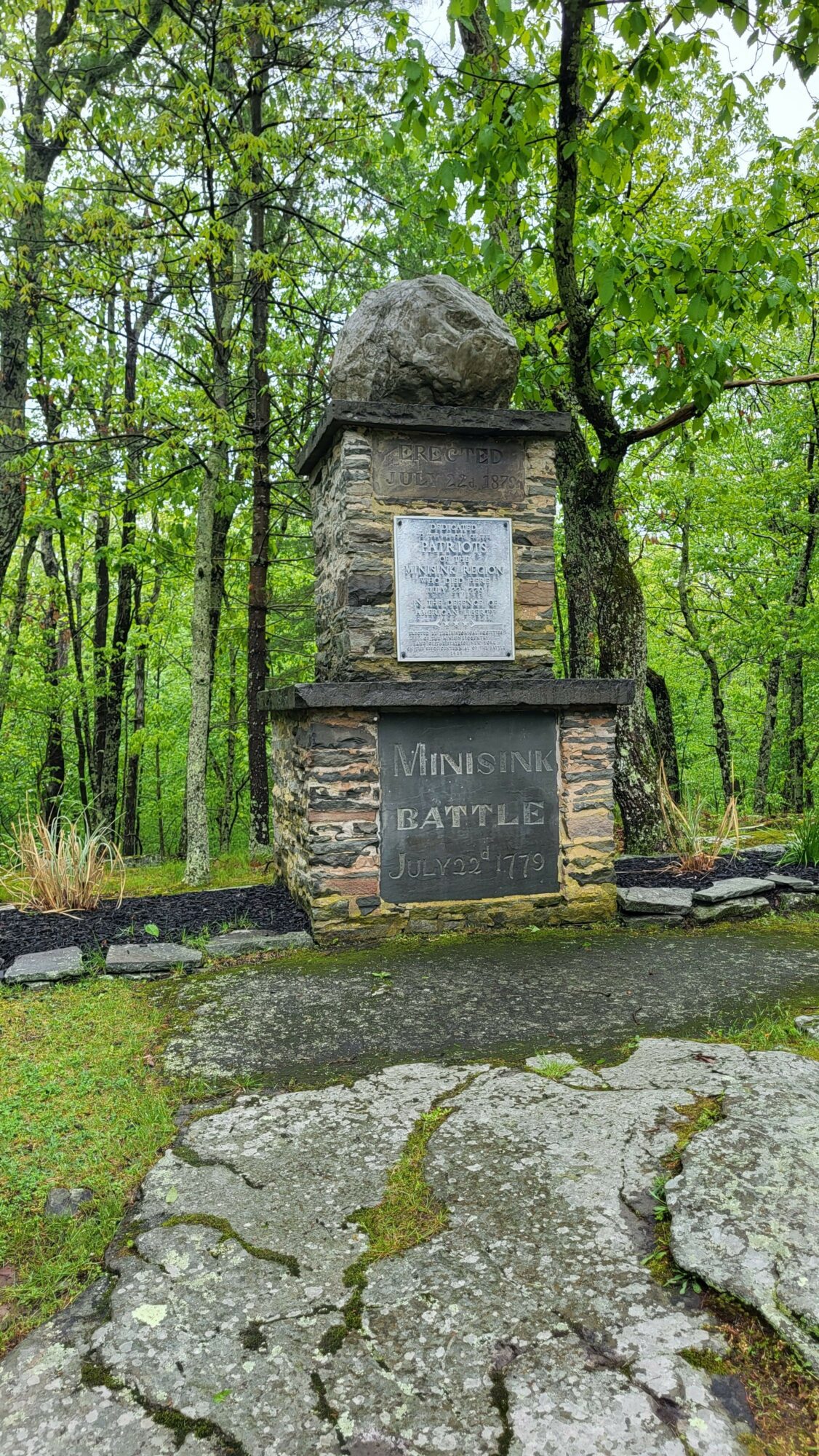 Stone monument with plaques in a green forested area, surrounded by trees and moss-covered rocks.