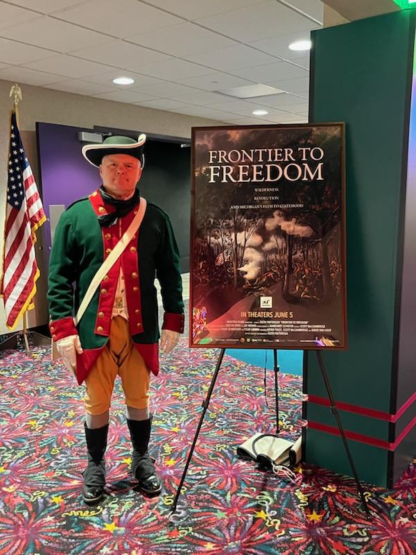 Person dressed as a historical soldier standing next to a poster titled 'Frontier to Freedom' in an indoor setting with an American flag in the background.