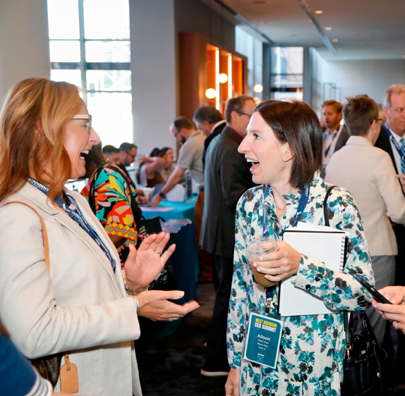 Two women are talking and smiling at a professional event, with other people in the background. One woman has red hair and glasses, the other has dark hair and is holding a glass and a notebook.