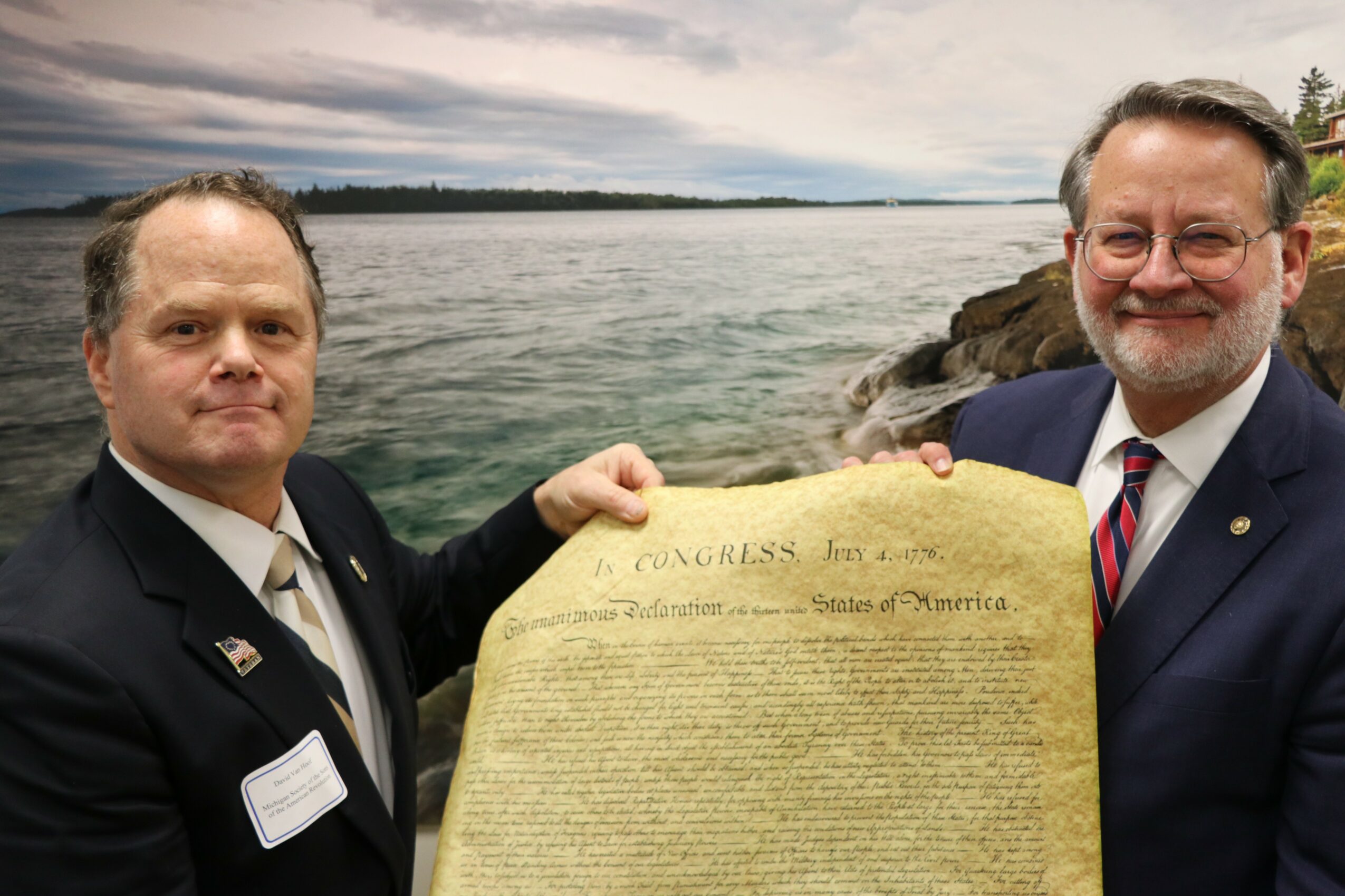 Two men in suits holding a large document outdoors near water, with a cloudy sky and distant shoreline in background.