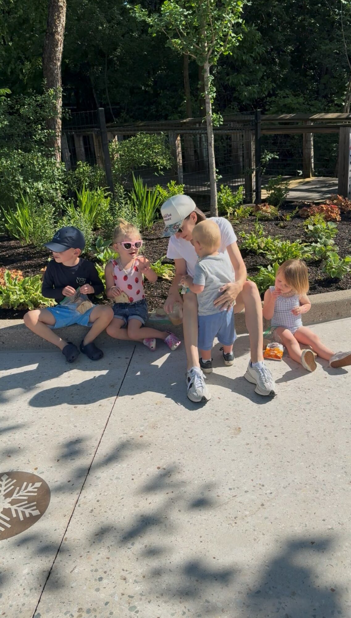 Four children sitting and standing on a sidewalk in a park, with trees and a fence in the background.