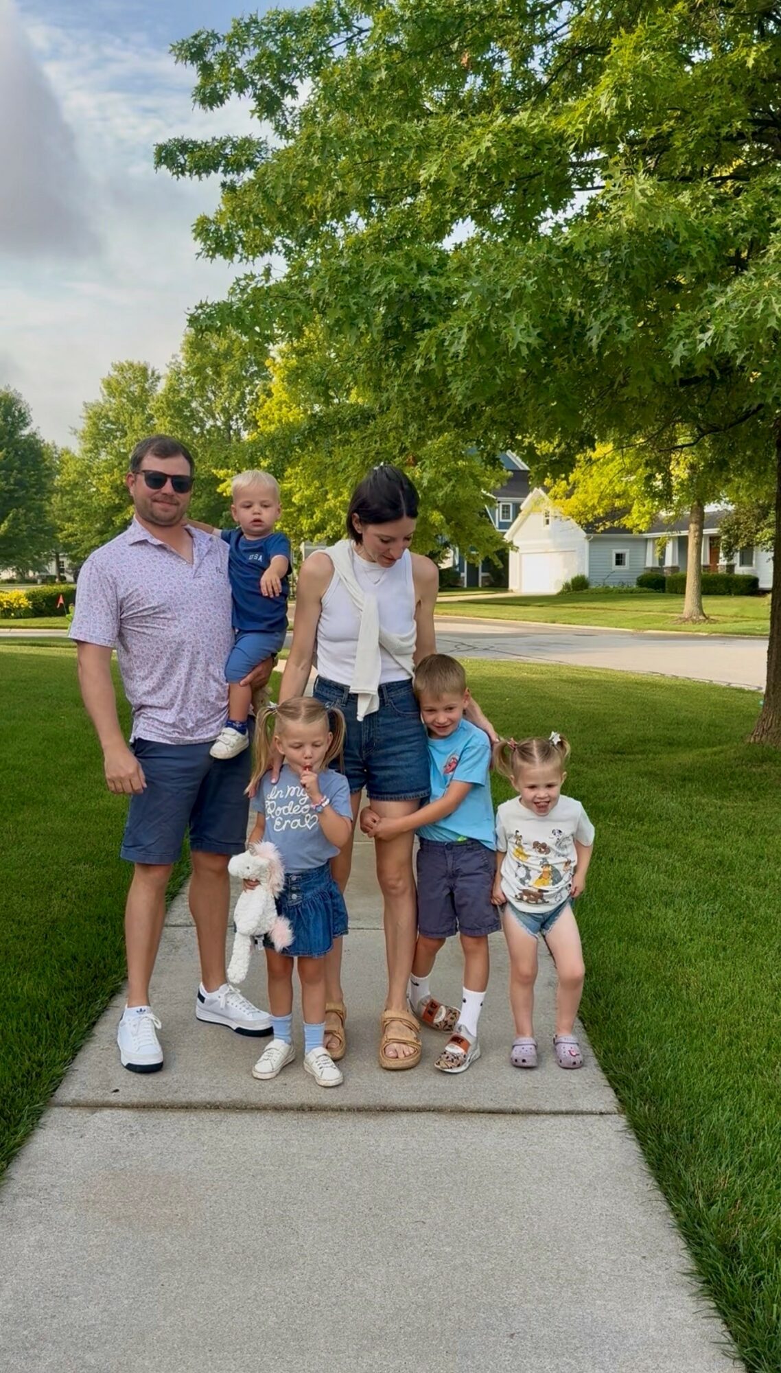 Family of five with two children and two adults walking on a sidewalk in a park, trees and houses in background.