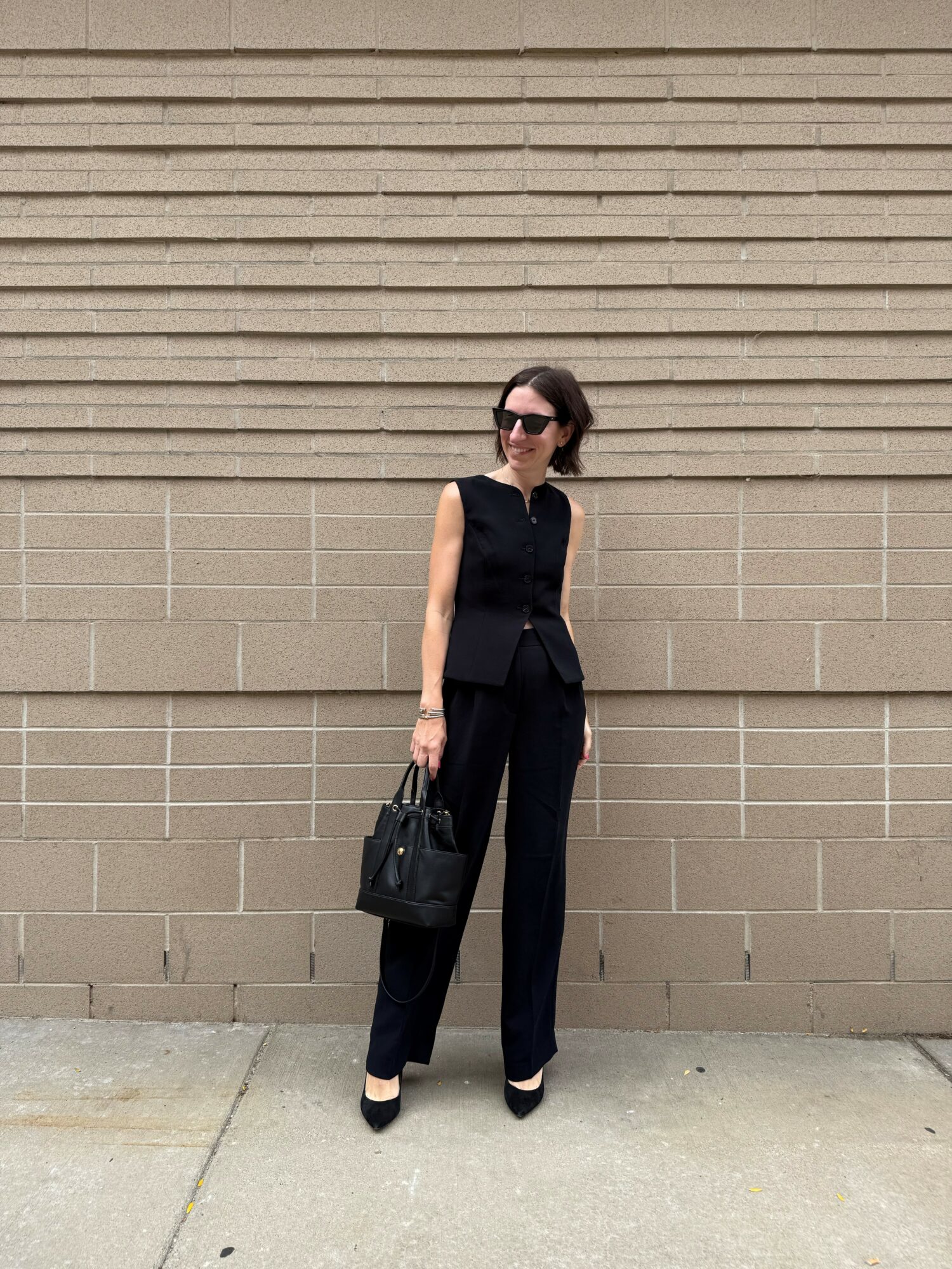 Woman in black sleeveless top and pants holding a black handbag, standing against a beige brick wall, wearing sunglasses.