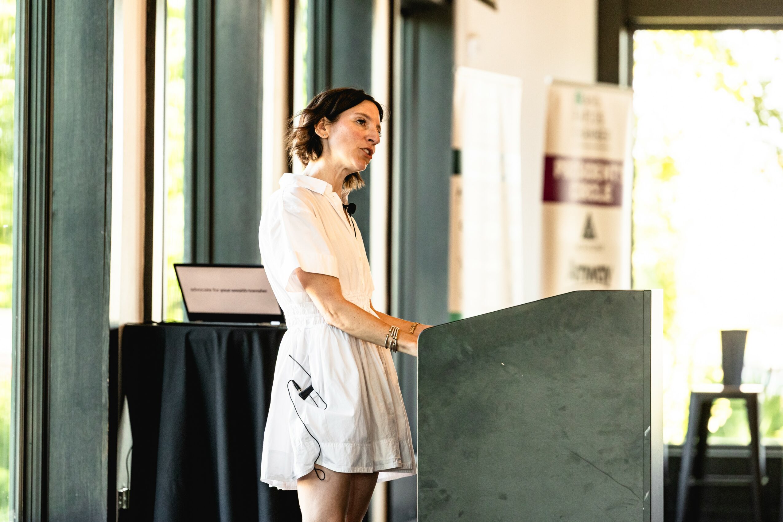 Woman in white dress standing at a podium in a room with large windows, sunlight streaming in.