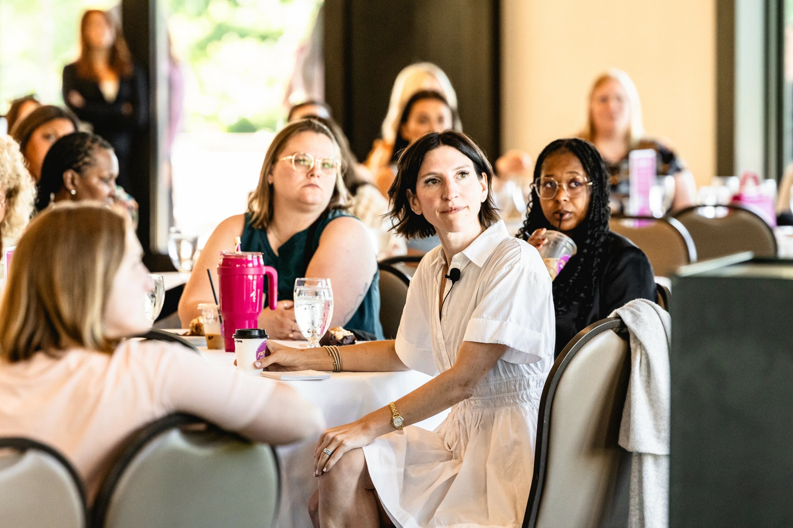 Group of women sitting at a table in a conference room, listening attentively, with drinks and notebooks in front.
