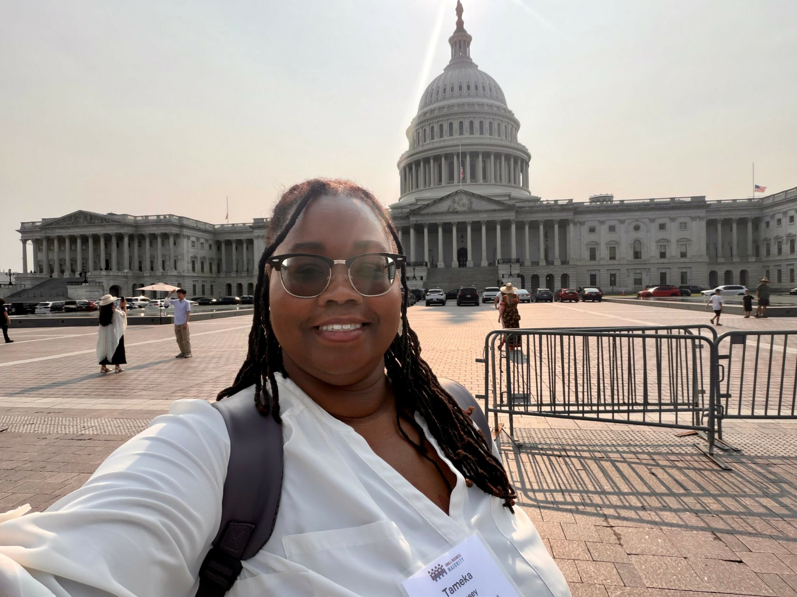 Person with glasses and a backpack taking a selfie in front of the U.S. Capitol building, sunny day.