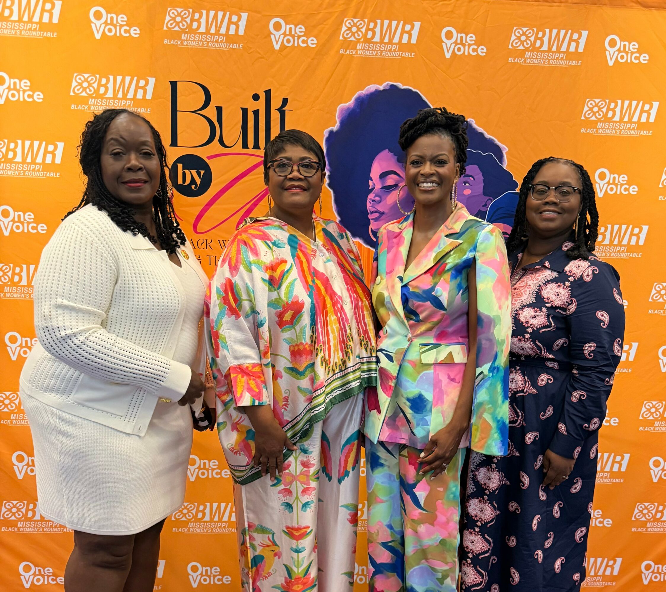 Four women stand together in front of an orange backdrop with logos and text, smiling at the camera.
