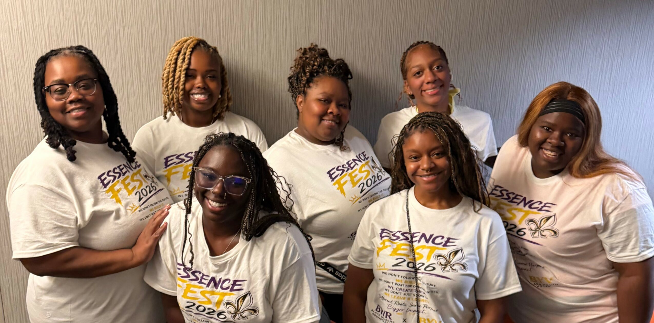 Group of eight women smiling, wearing white T-shirts with yellow and blue text, standing against a plain wall.