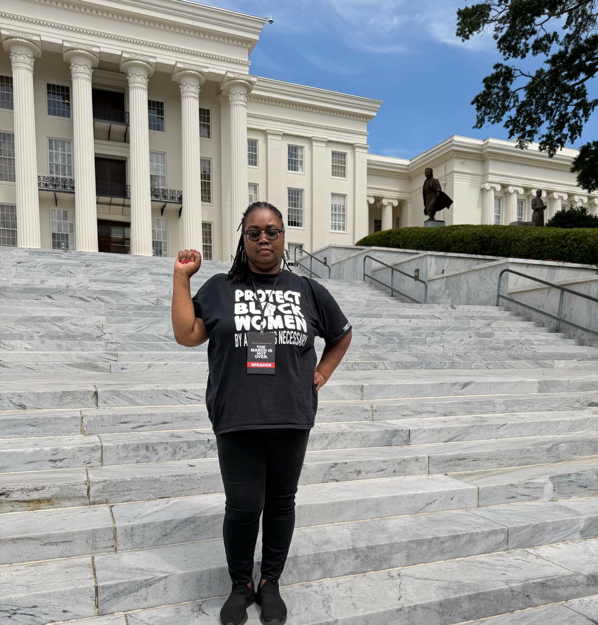 Person standing on marble stairs in front of a white building with columns, raising a fist, wearing a black t-shirt and sunglasses.