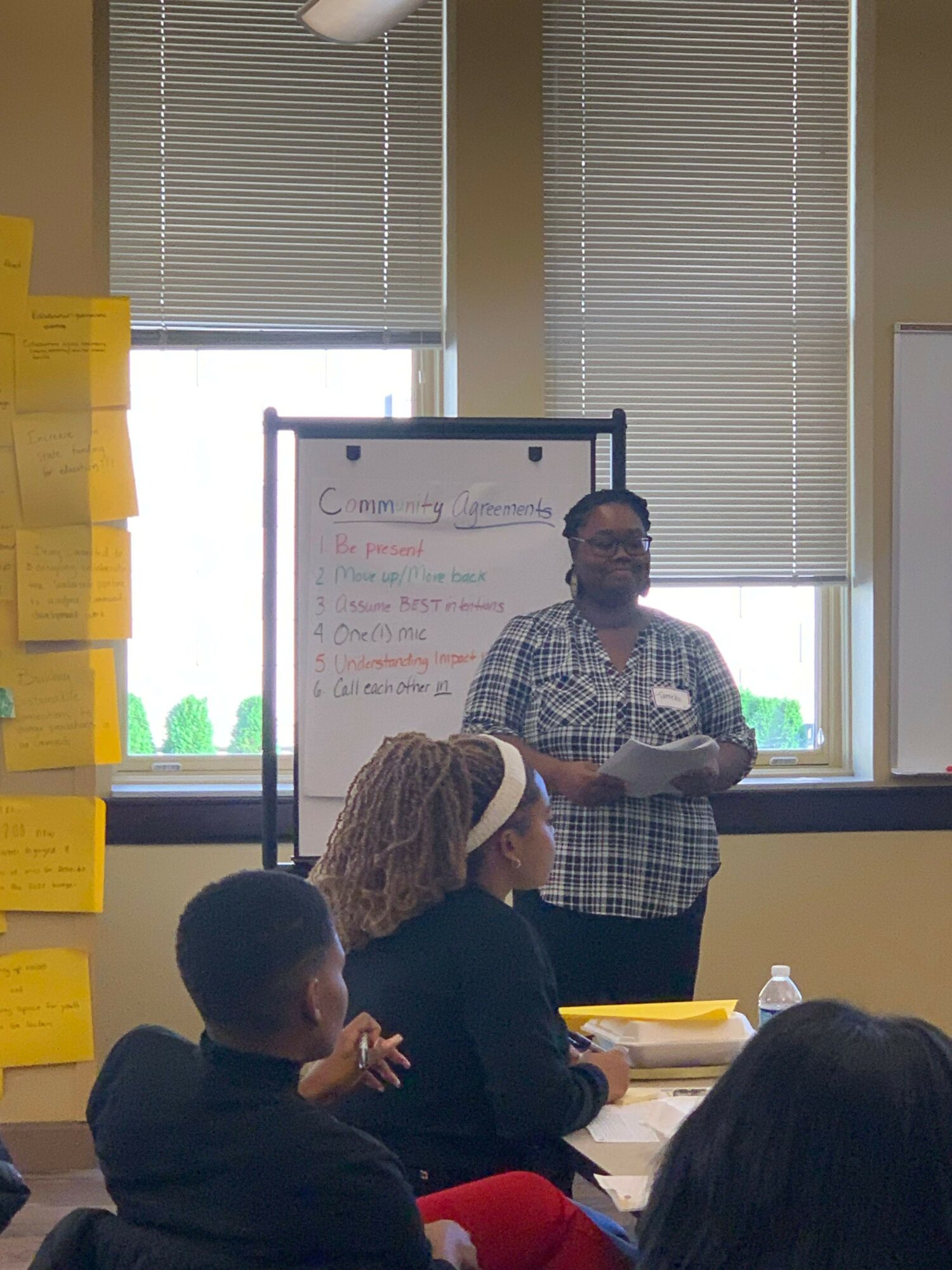 Person standing in front of a whiteboard, presenting to a group in a room with windows and blinds.
