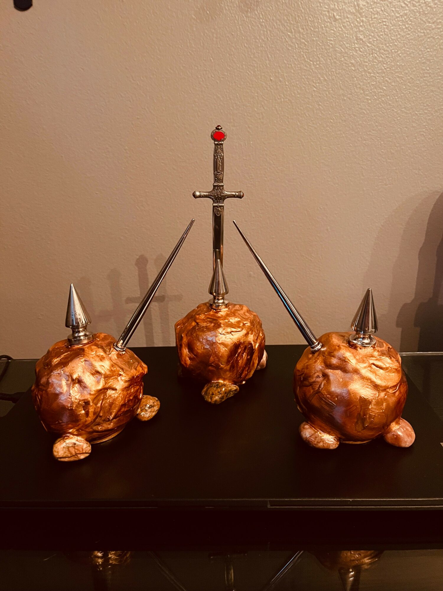Three decorative objects resembling pigs with metallic spikes and a sword, arranged on a black surface against a beige wall.