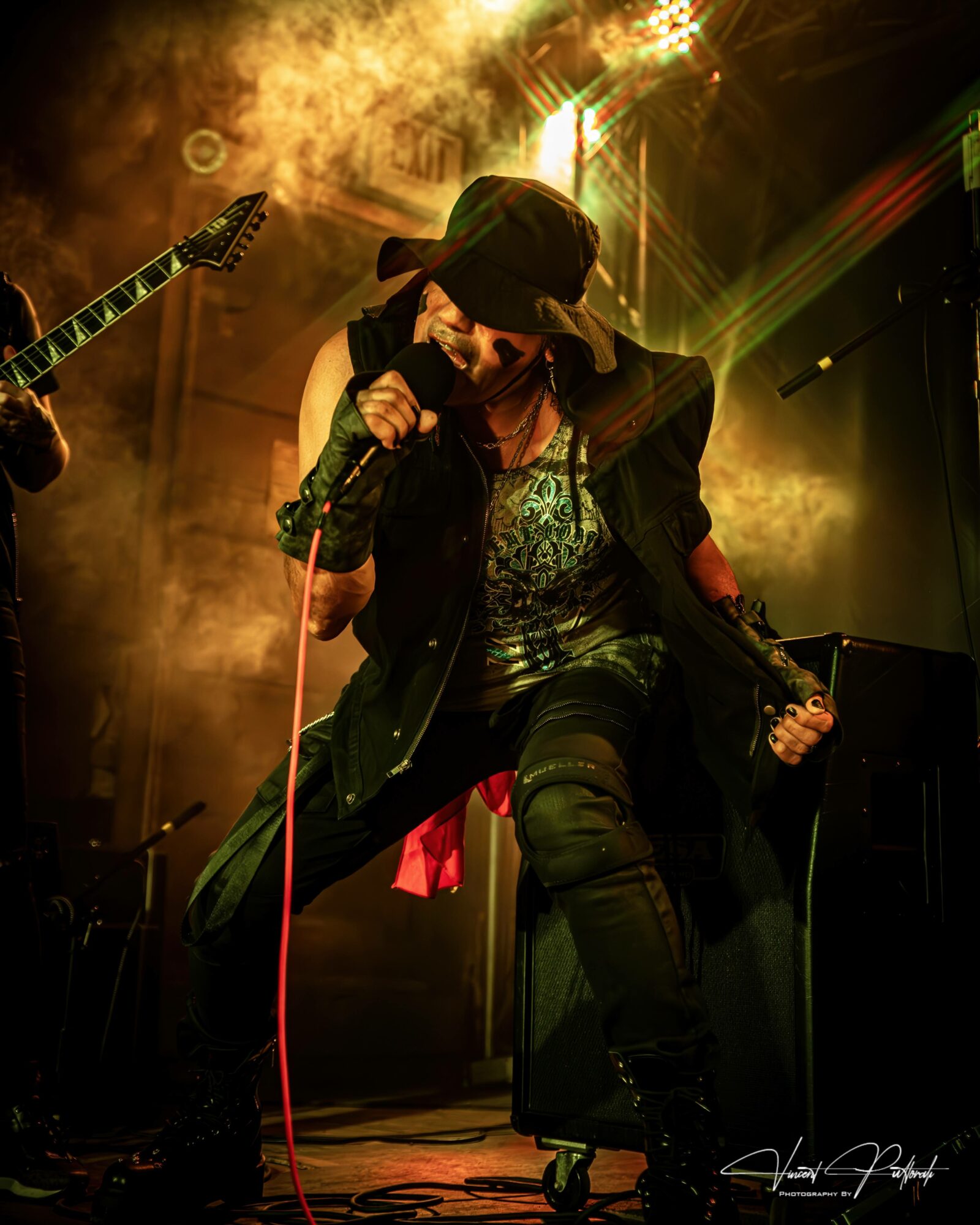 Performer singing into microphone on stage with colorful lights and smoke effects, wearing a cap and dark clothing.