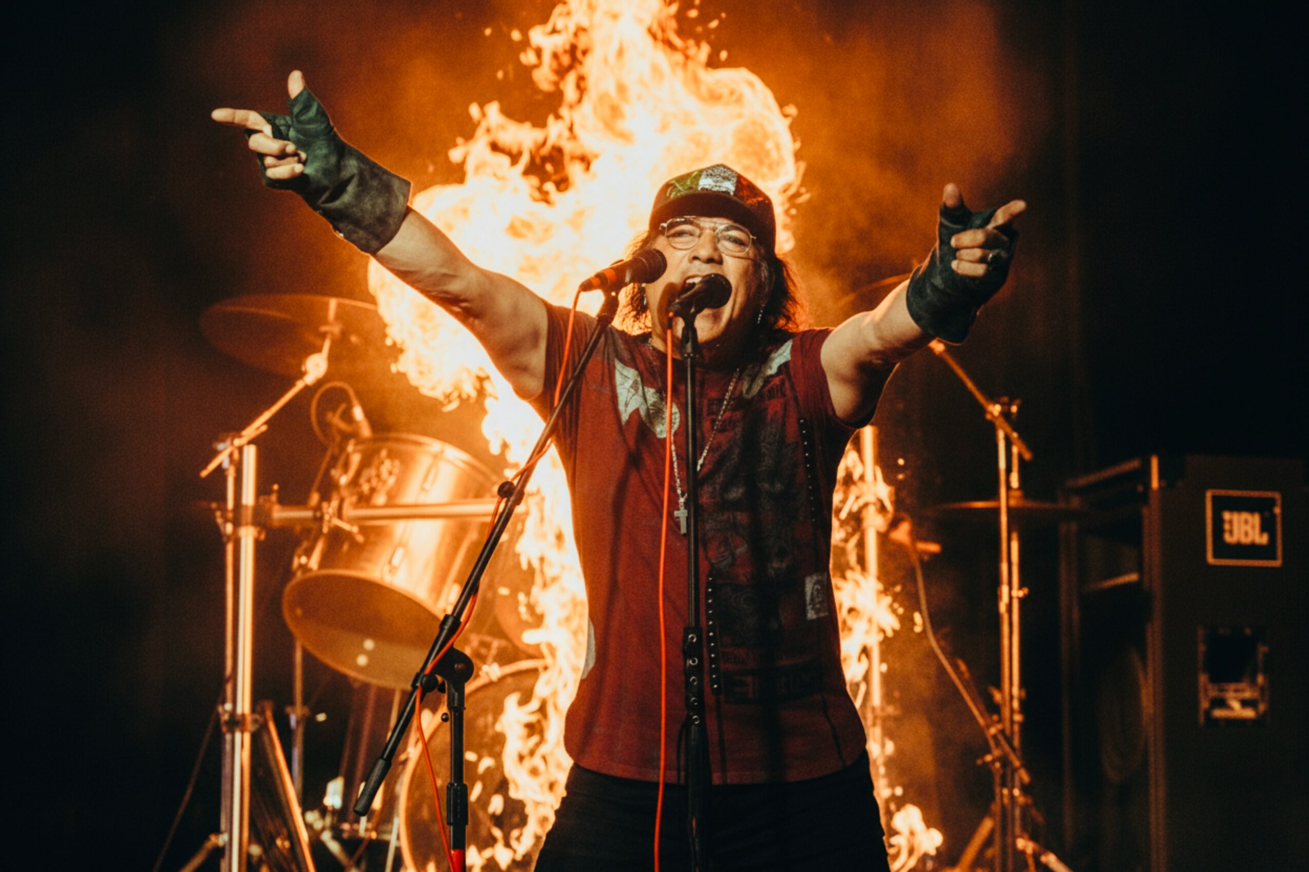 Performer singing with arms outstretched in front of flames and drums, on stage with musical equipment, expressive pose.