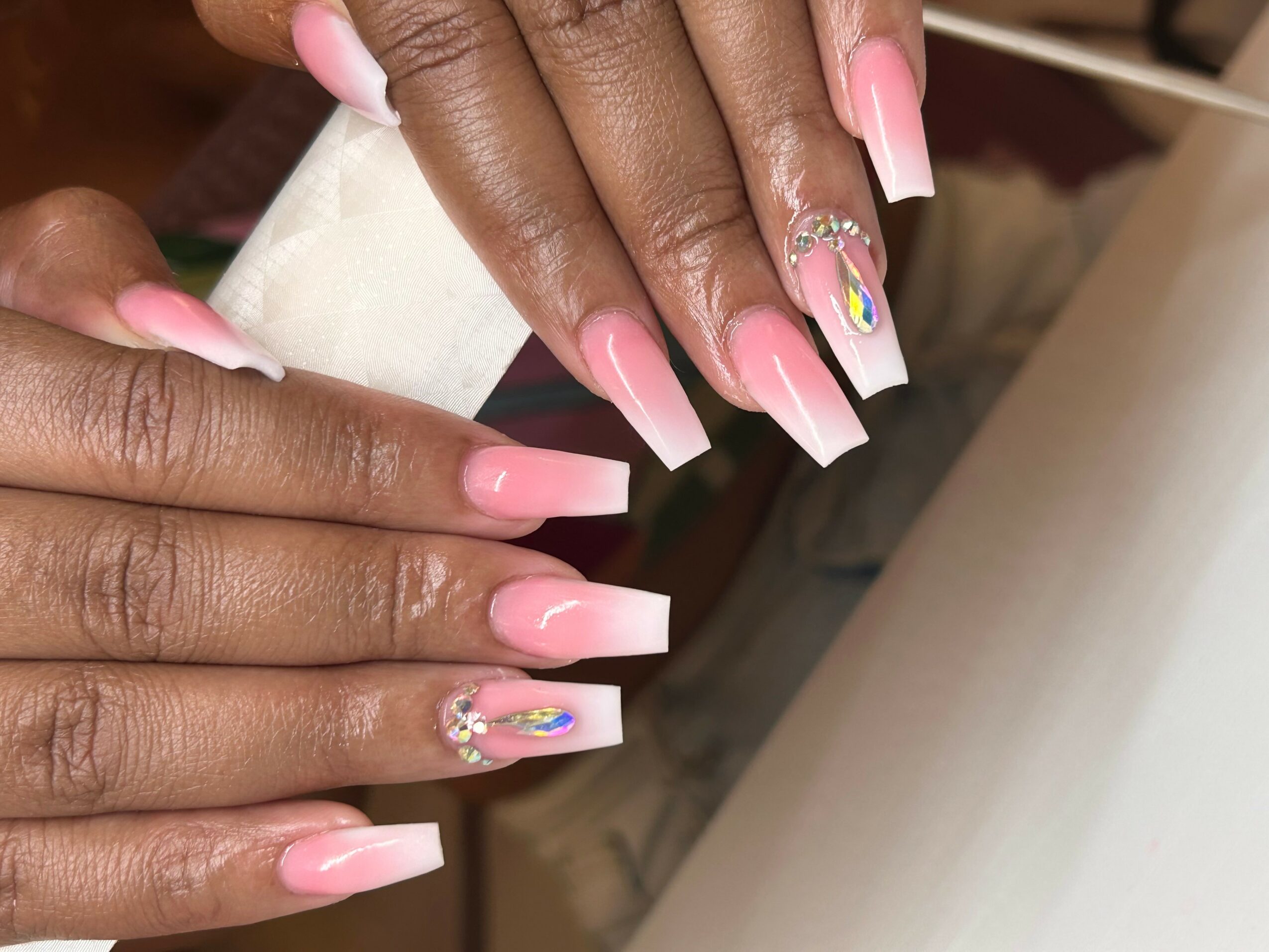 Close-up of hands with long, pink, square-shaped nails, some with rhinestone and rainbow designs, on a white surface.