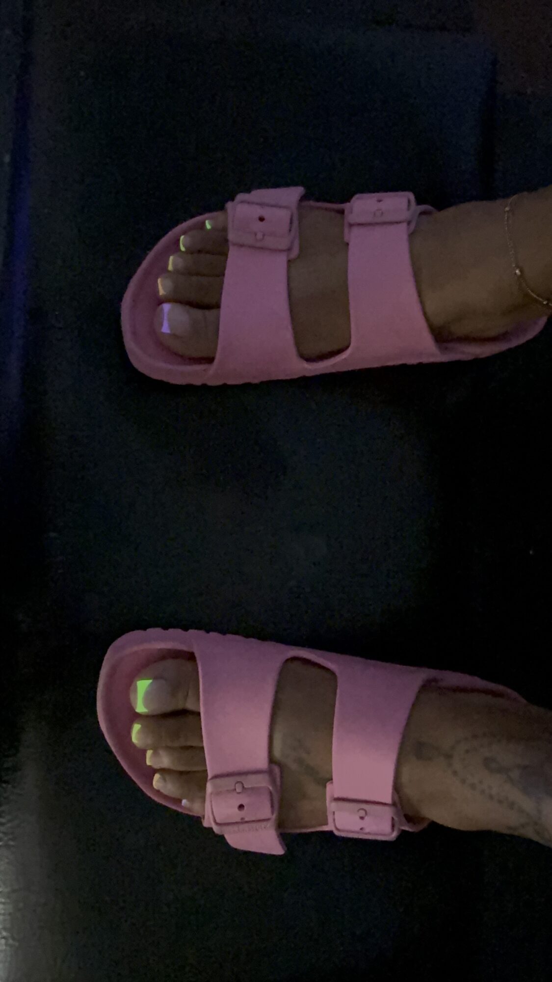 Person wearing pink sandals with two straps, toes visible, on a dark surface.