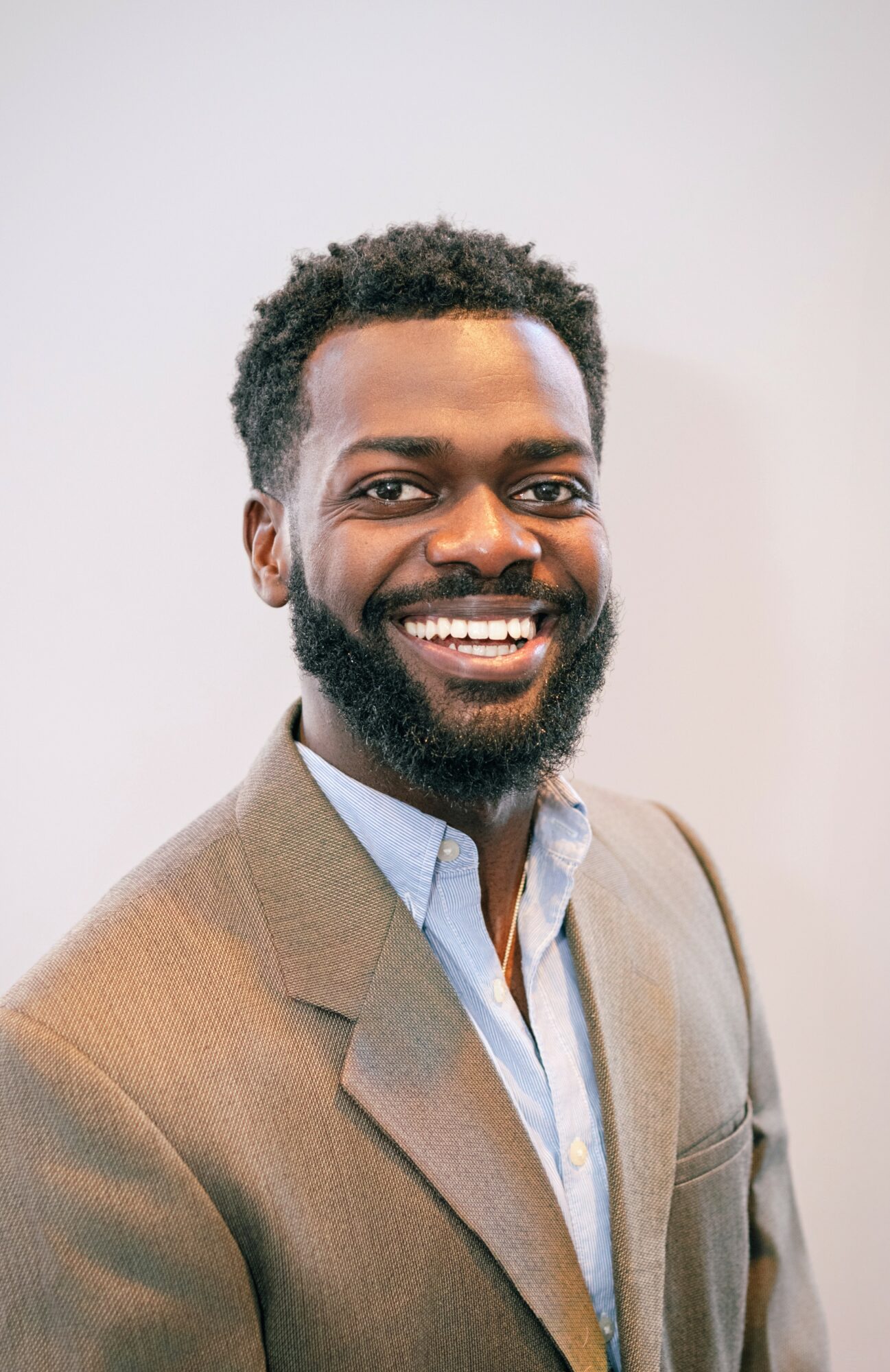 Smiling man with a beard and curly hair wearing a beige blazer and light blue shirt, standing against a plain background.