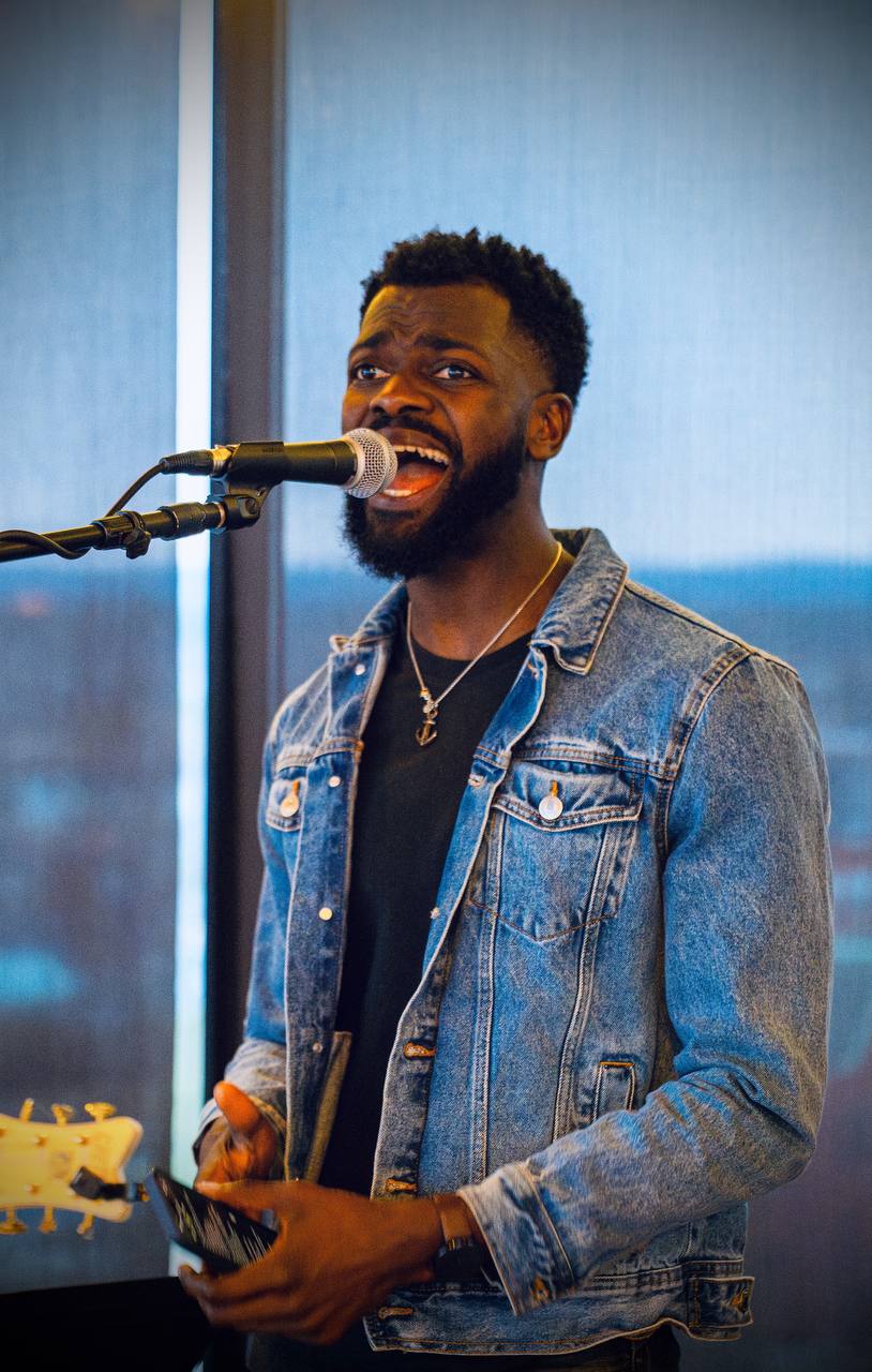 Man singing into microphone, wearing denim jacket and black shirt, holding a guitar, smiling, indoors with blue background.