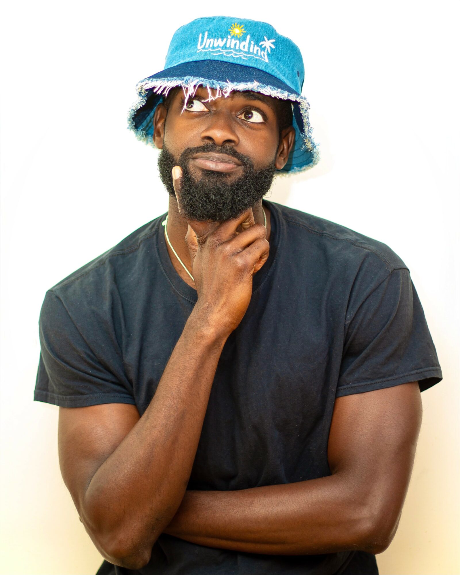 Man with a beard wearing a blue bucket hat and black t-shirt, touching his chin thoughtfully.