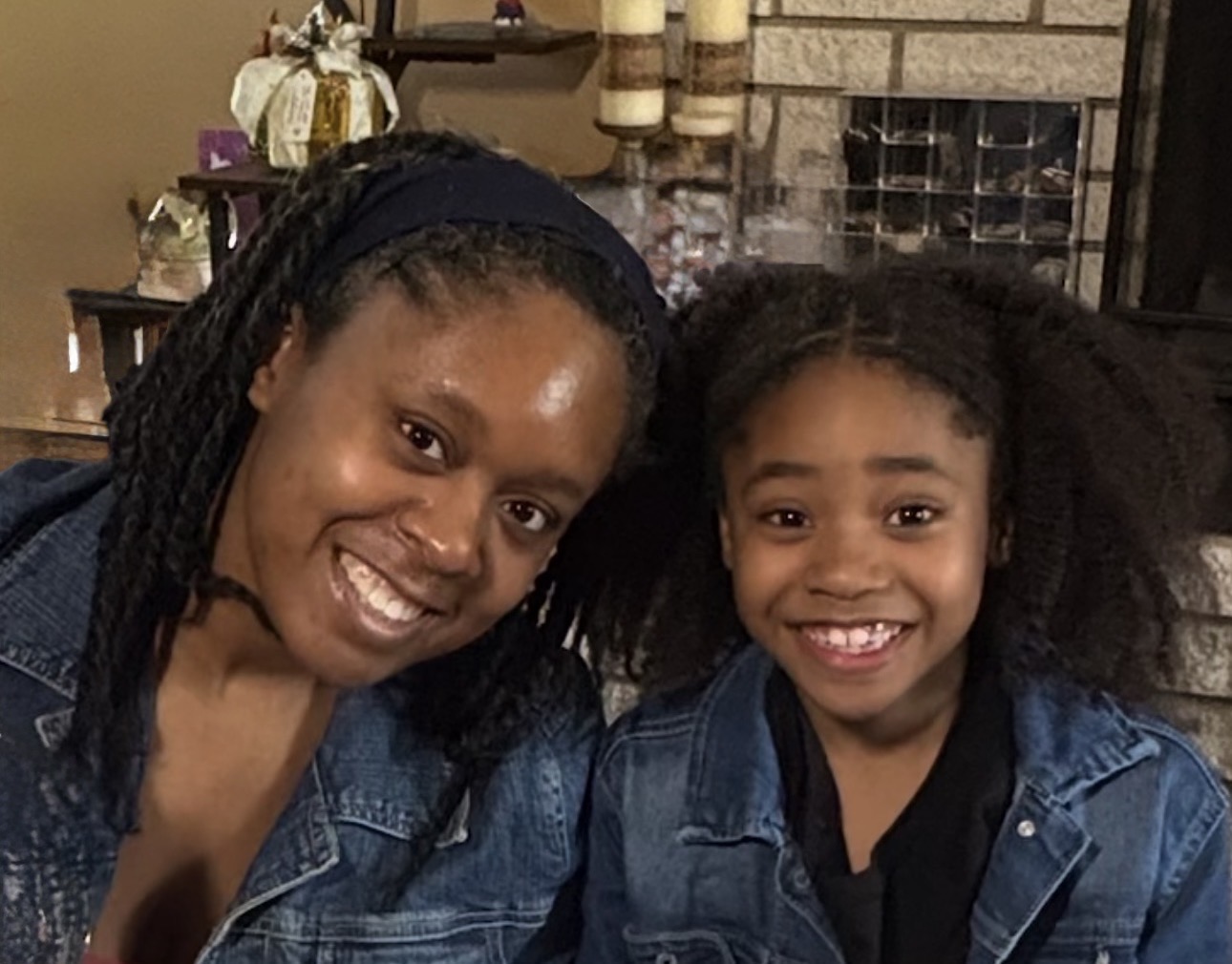 Woman and girl smiling, both wearing denim jackets, sitting indoors with a brick wall and decorations in the background.