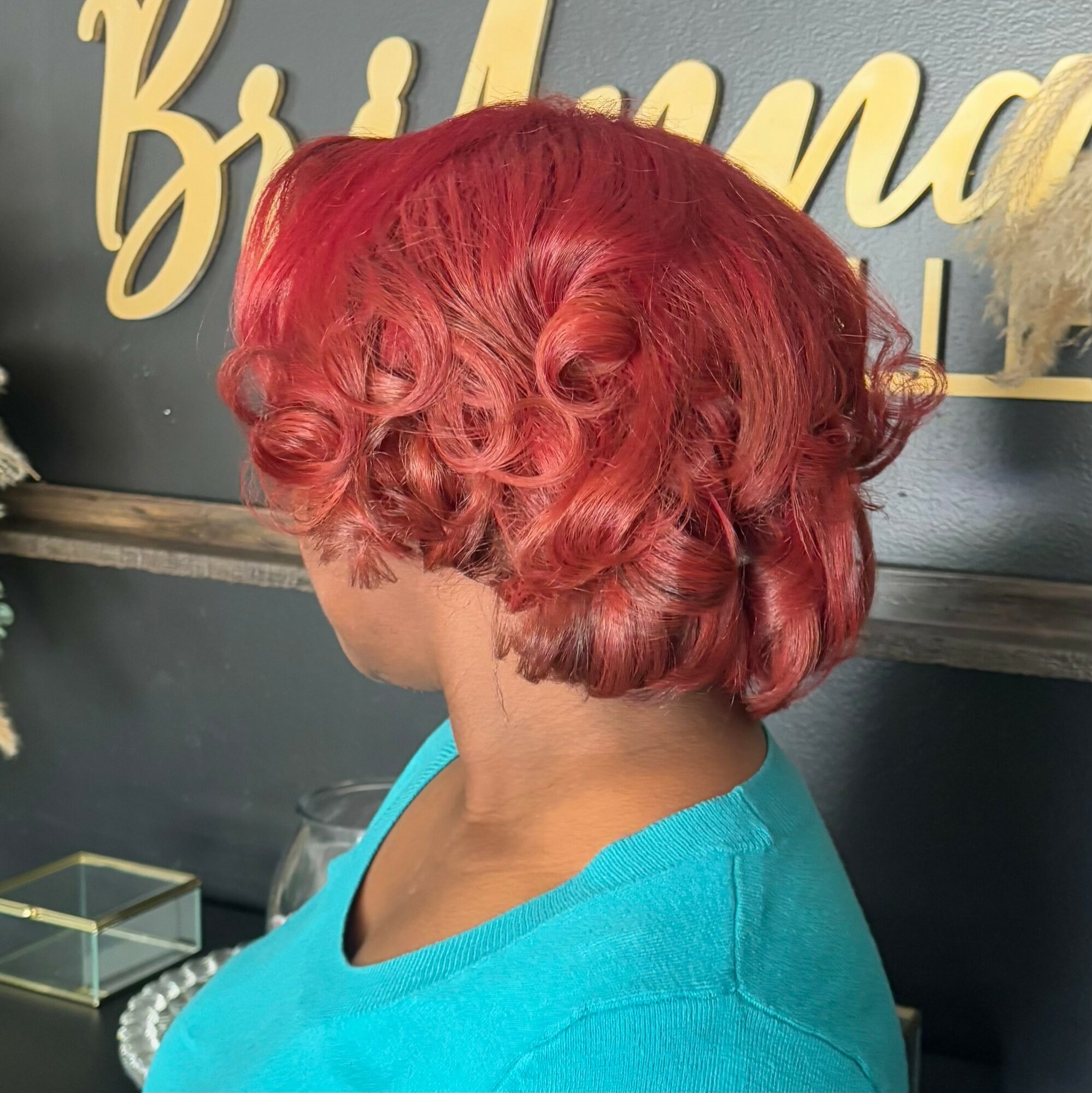 Person with short, curly red hair wearing a blue top, facing away from the camera, in a salon setting.