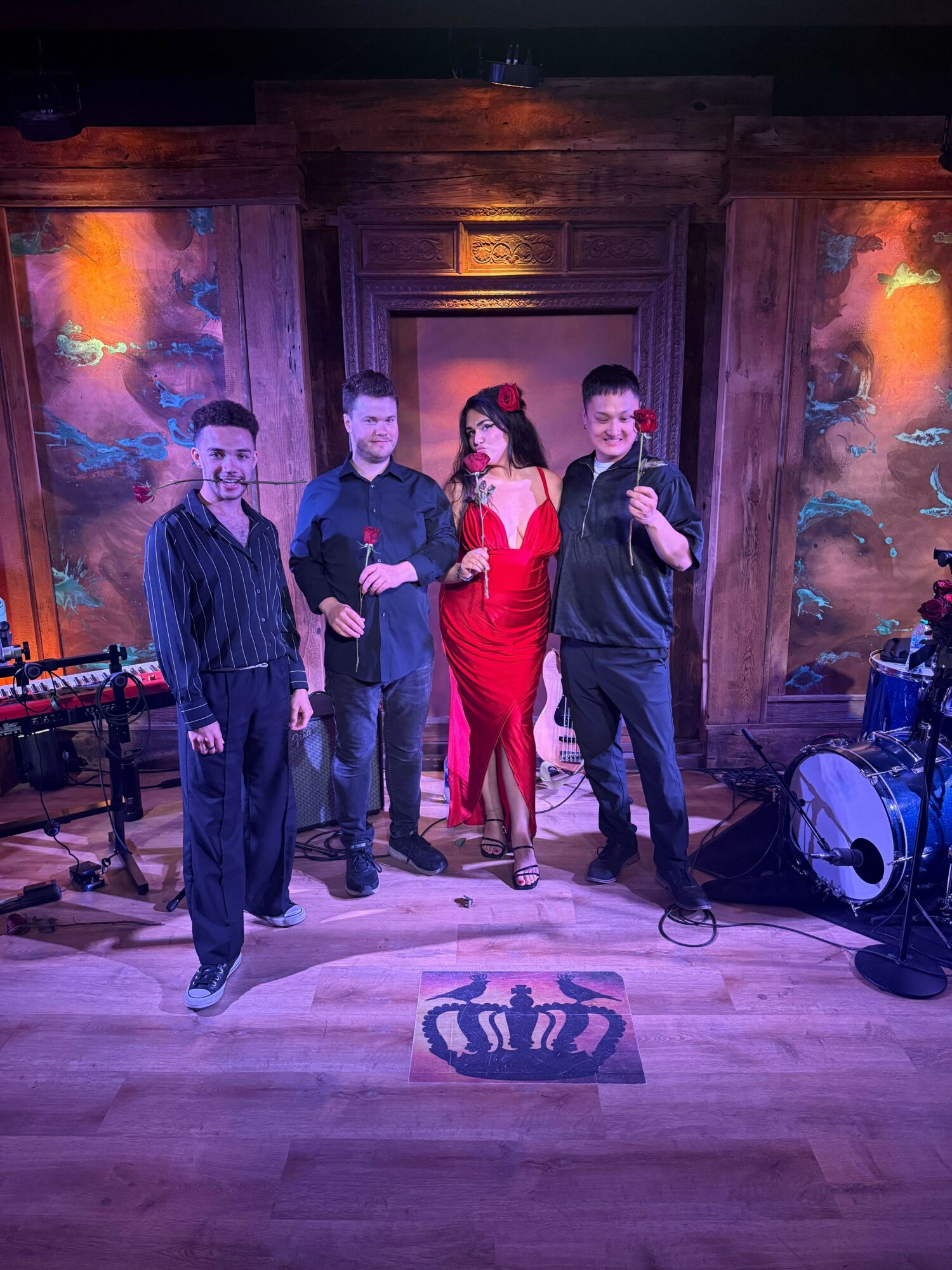 Four people stand in a room with wooden walls and colorful artwork, posing for a photo. One person wears a red dress, others in dark clothing.