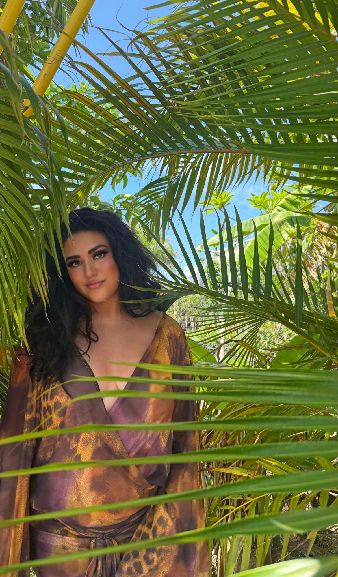 Woman with dark hair and makeup among green palm leaves, wearing a colorful top, in a tropical setting.