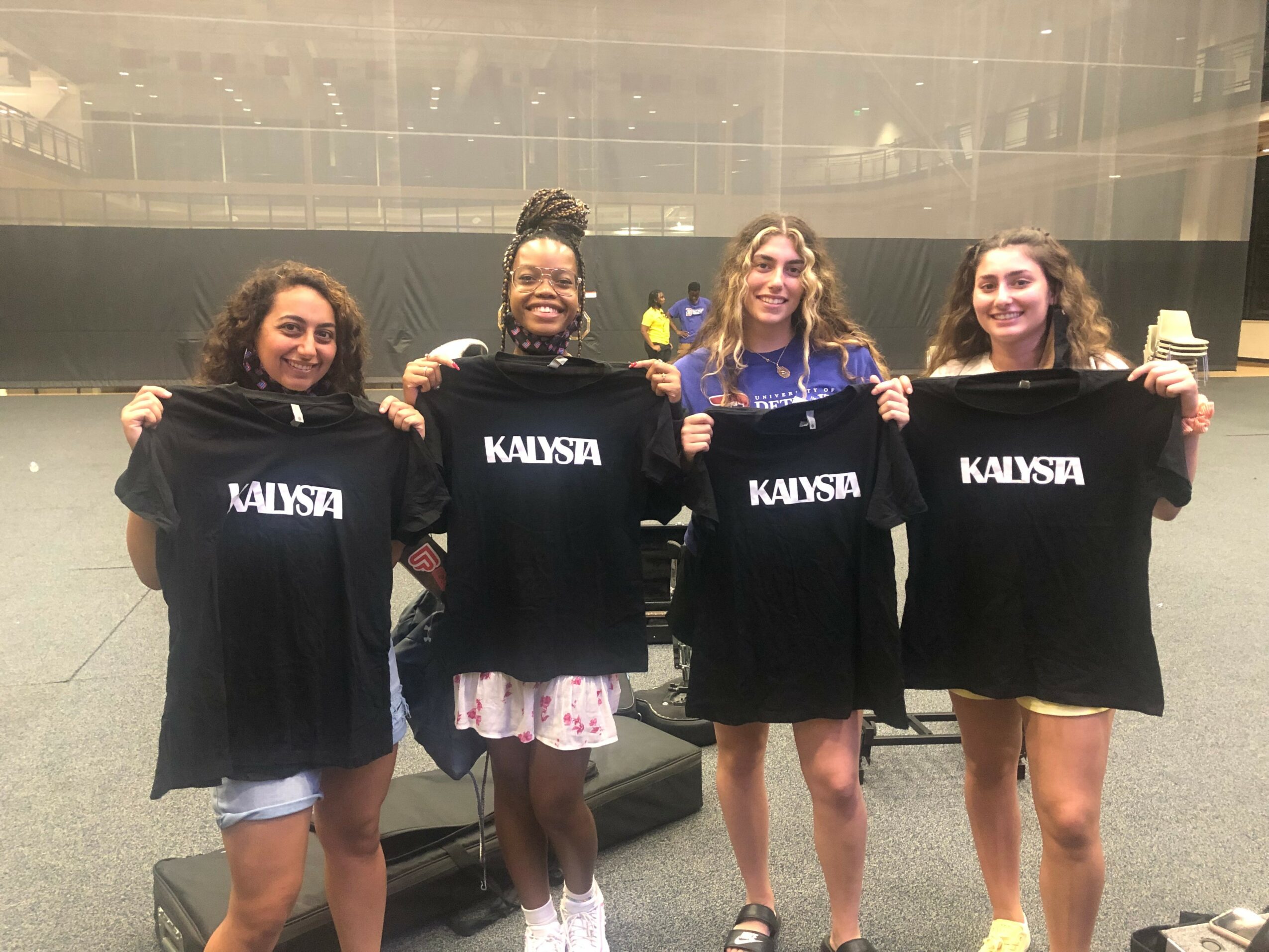 Four young women holding black T-shirts with white text, standing indoors in front of a glass wall.
