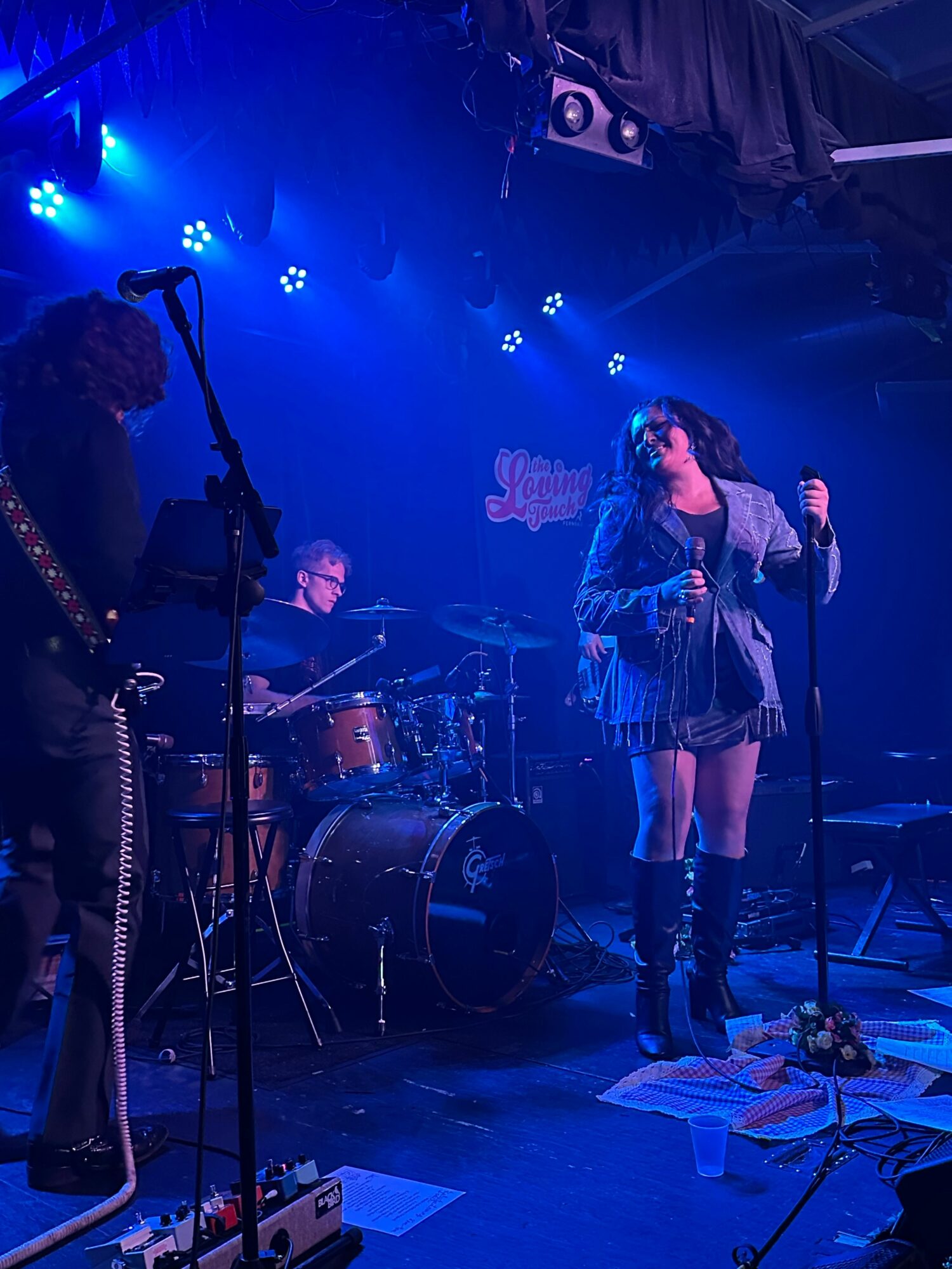 Two people perform on stage with musical instruments, blue stage lighting, and equipment visible, in a live music setting.