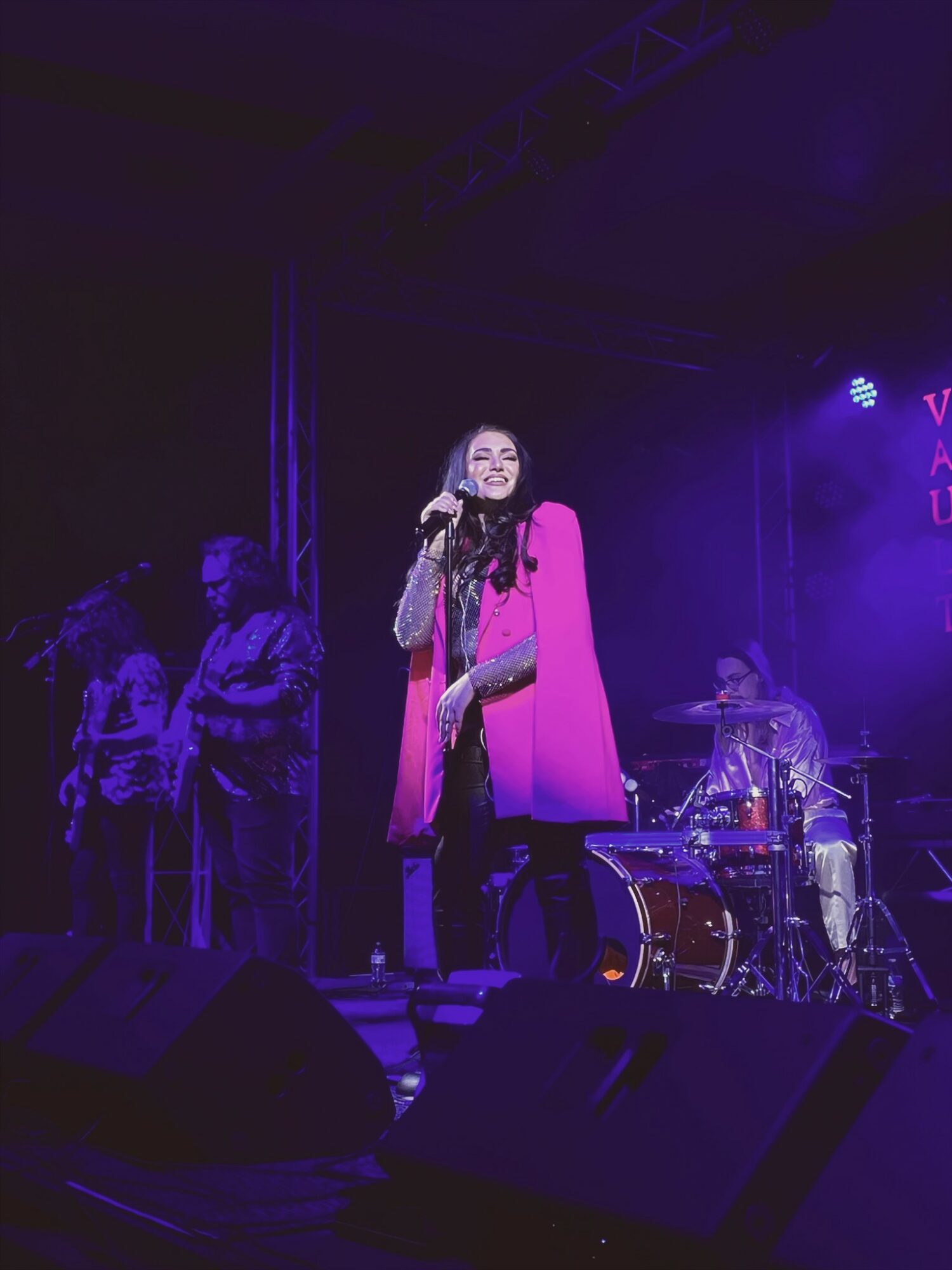 Performer singing on stage with purple lighting, surrounded by band members and audience, in a concert setting.