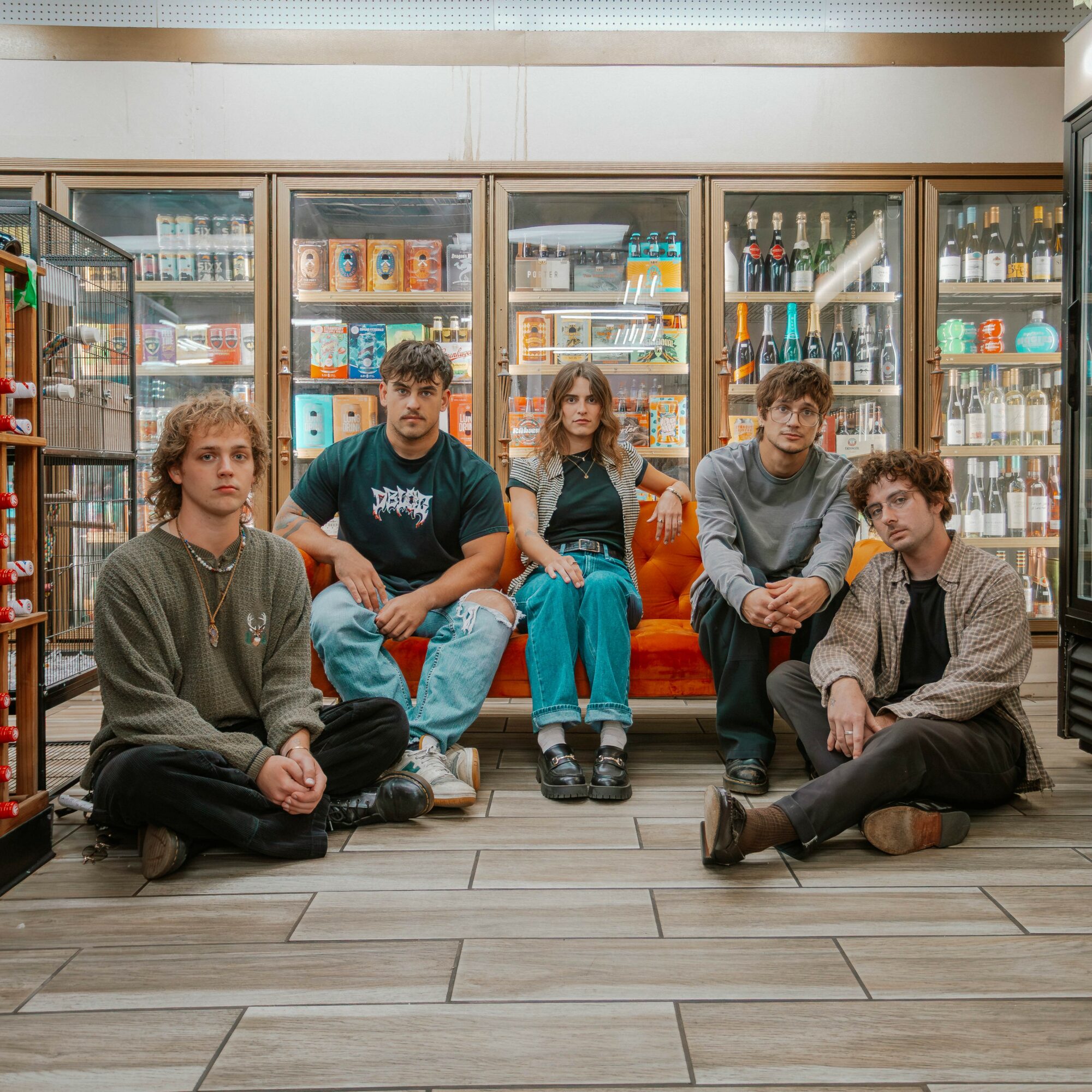 Five young people sit and kneel on the floor in front of a glass display case filled with various products, in a store. 20 words.