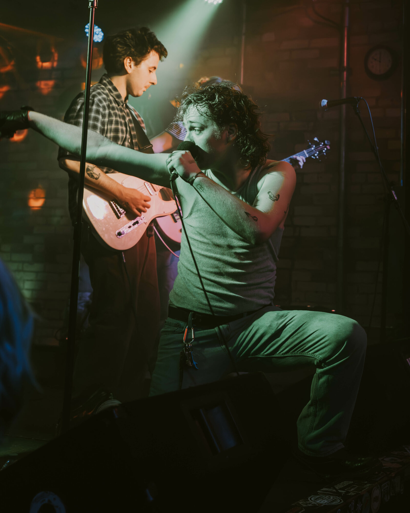 Two musicians perform on stage, one playing guitar and the other singing into a microphone, with stage lighting and brick wall background.