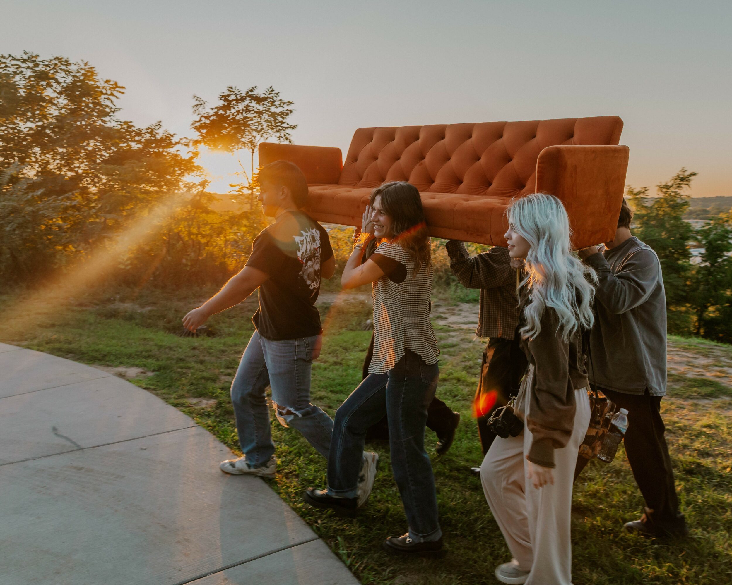 Group of people carrying a large orange sofa outdoors during sunset, walking on a path with trees in the background.