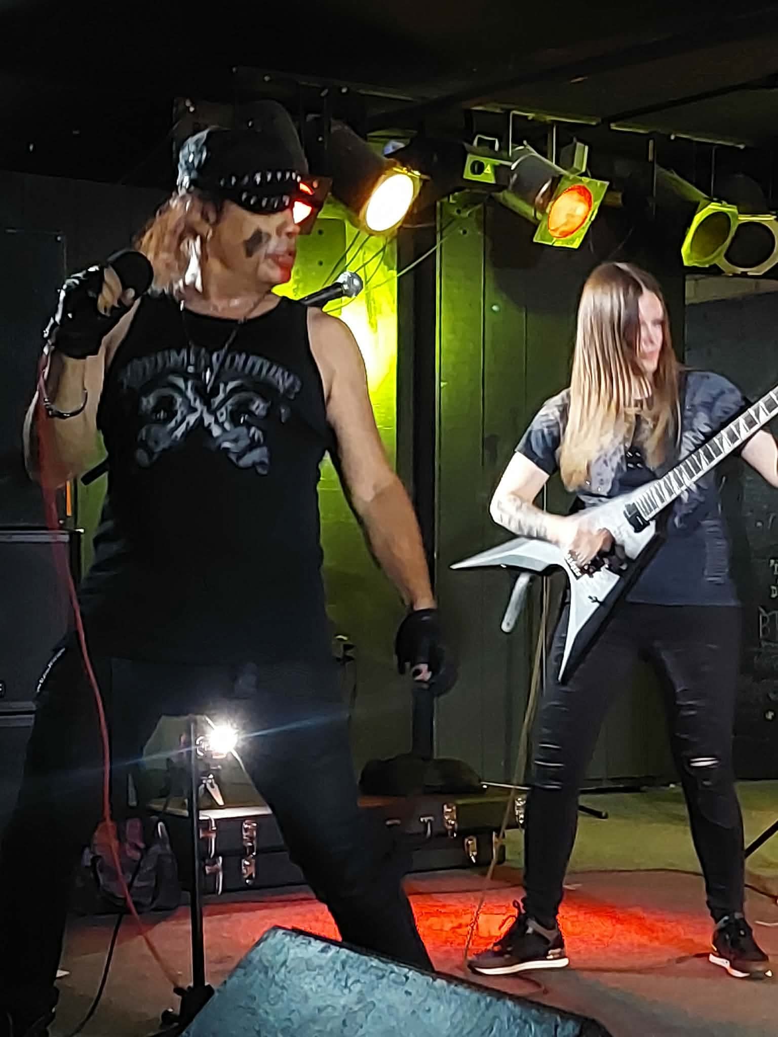 Two women perform on stage with musical instruments, stage lights illuminate them, one singing and the other playing guitar.