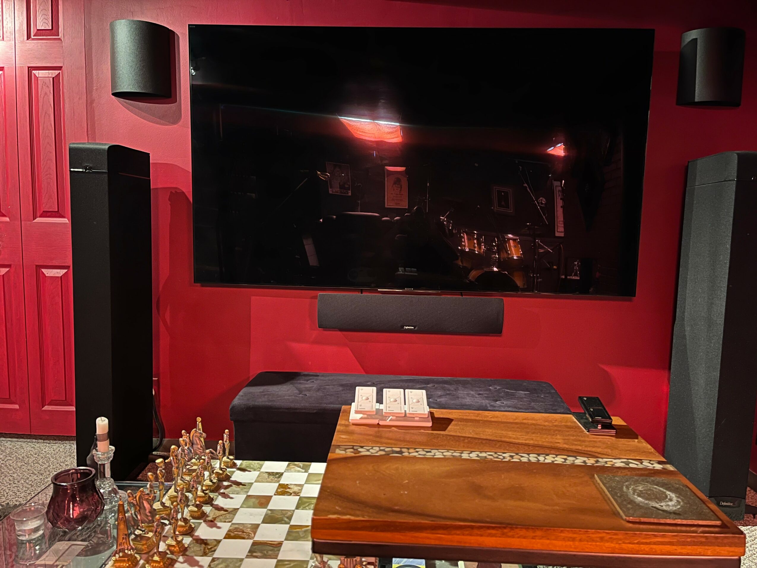 Living room with a large TV mounted on a red wall, surrounded by black speakers, a wooden table, and a black bench.