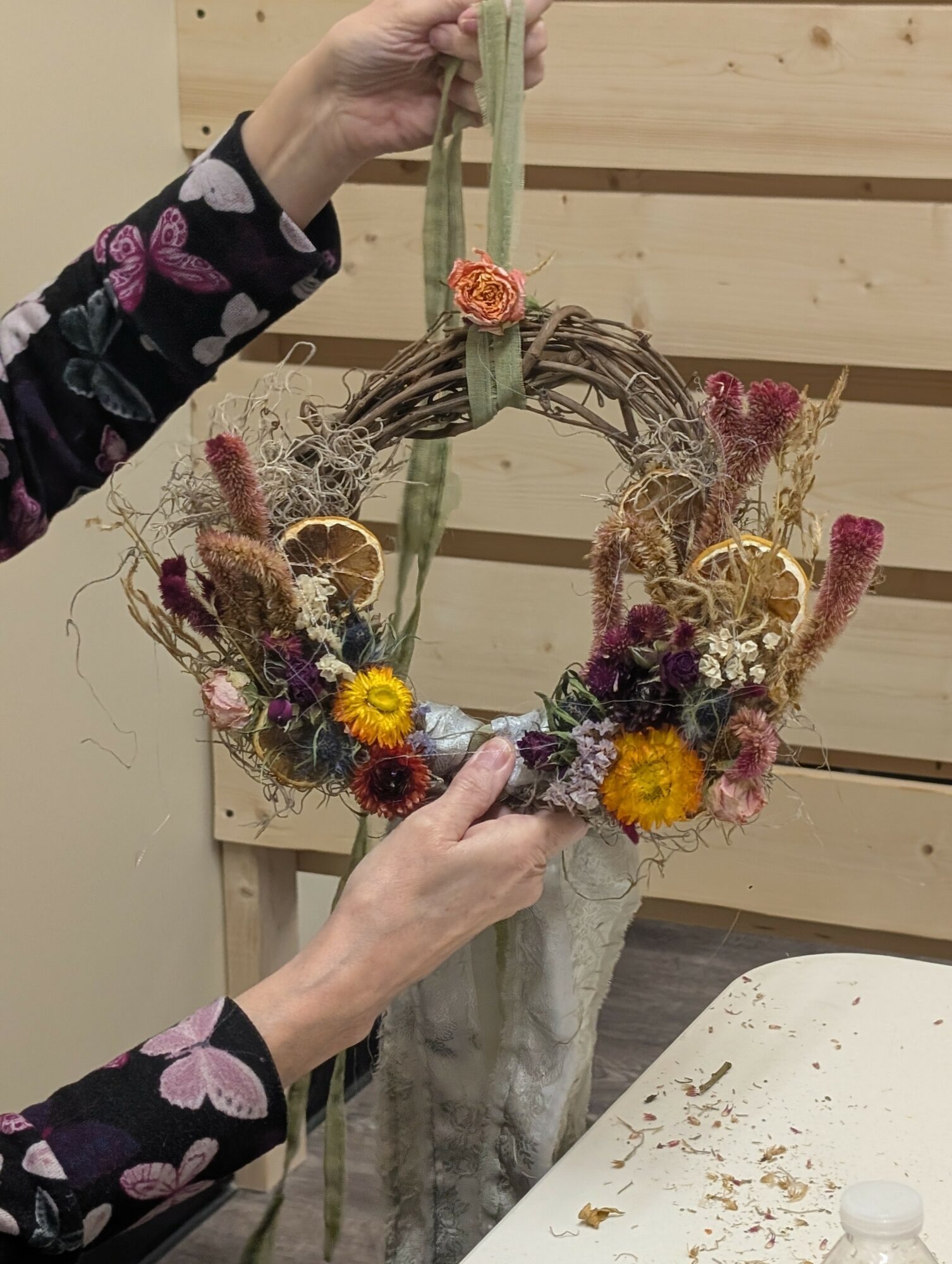 Person arranging a floral wreath with dried flowers and yellow accents, holding it in front of a wooden background.