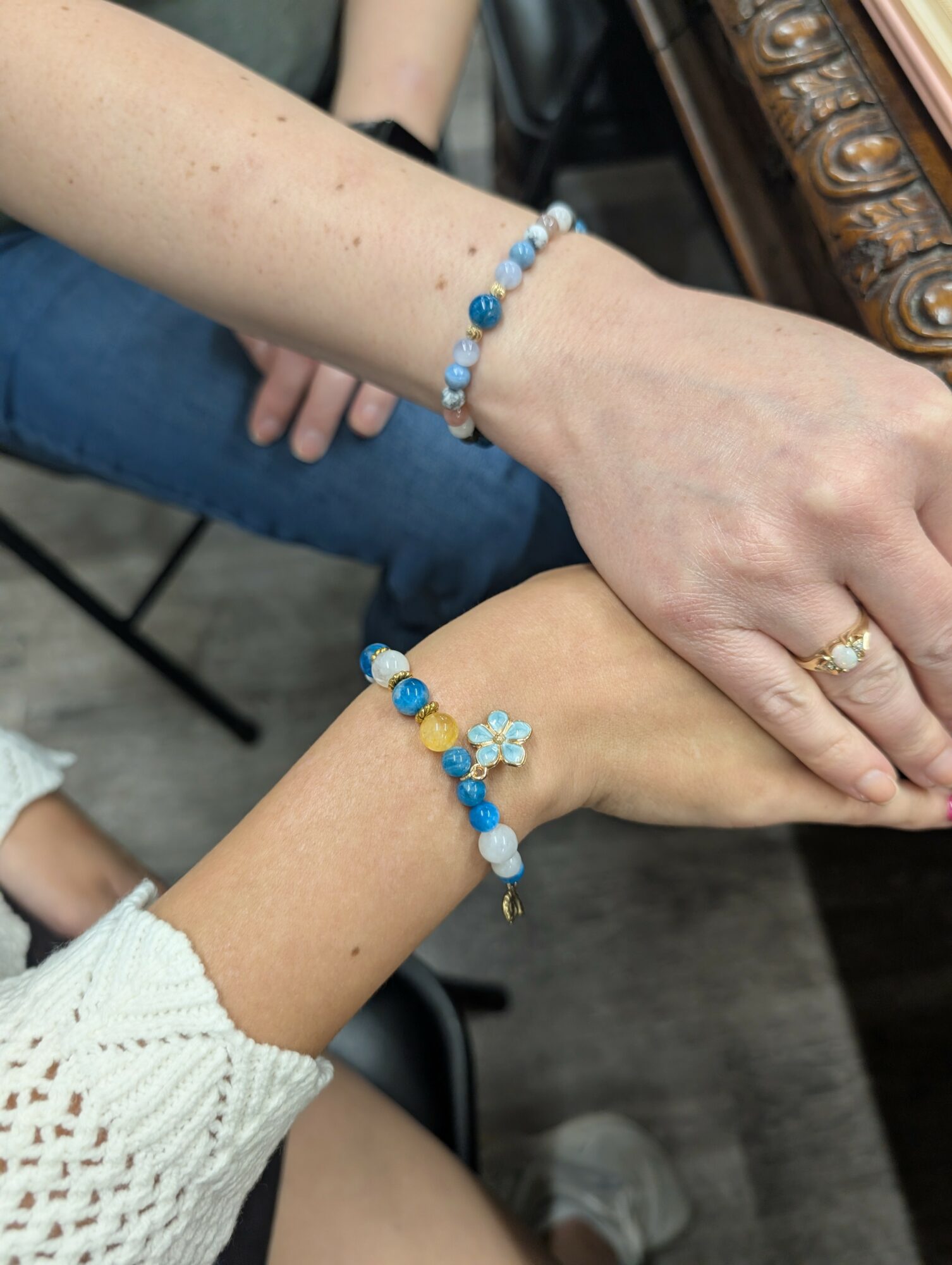 Two hands with matching blue and white beaded bracelets, one with a ring, resting on a wooden surface.