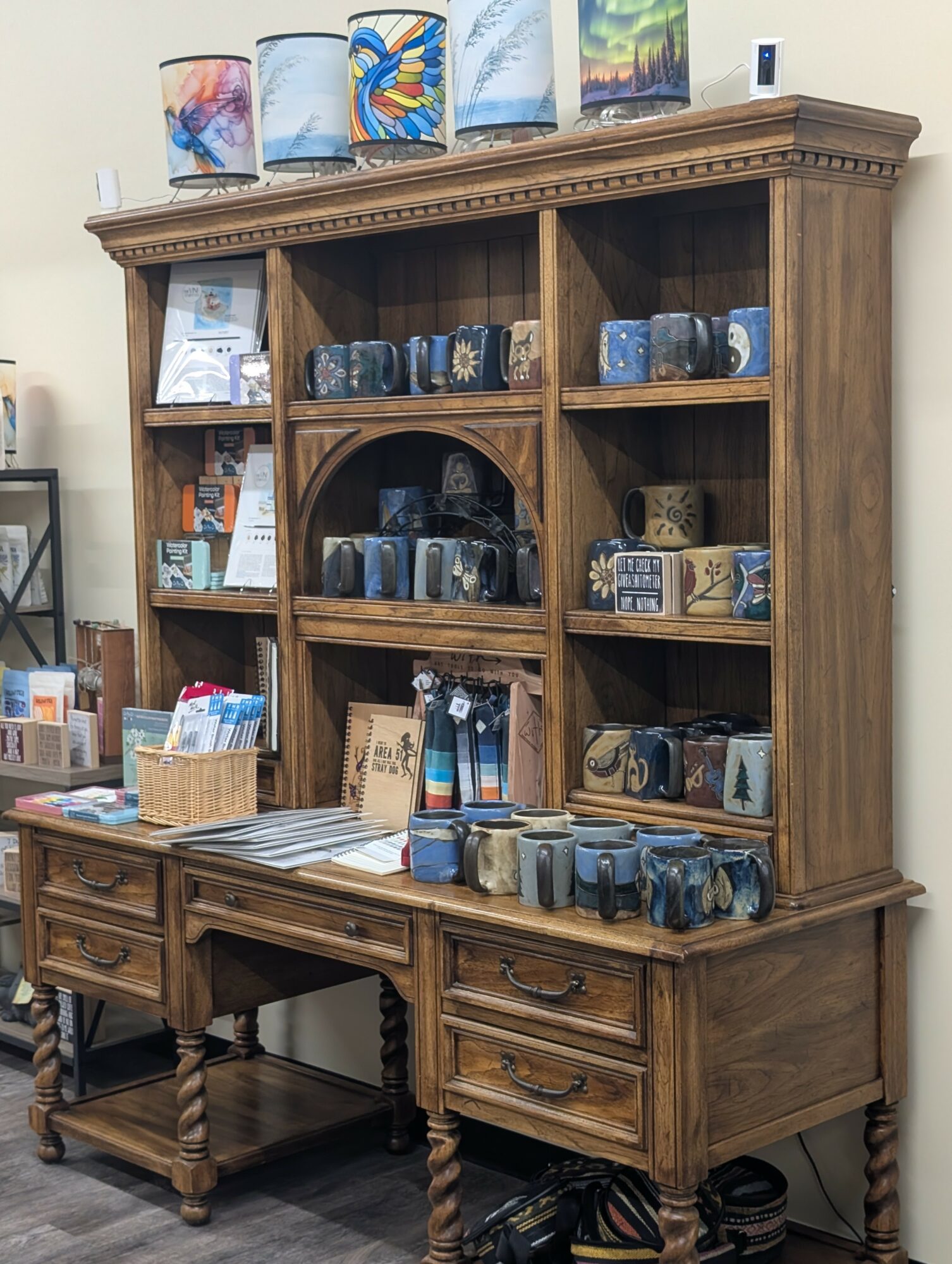 Wooden hutch with shelves filled with mugs, cups, and decorative items, and a desk with drawers below, in a store setting.