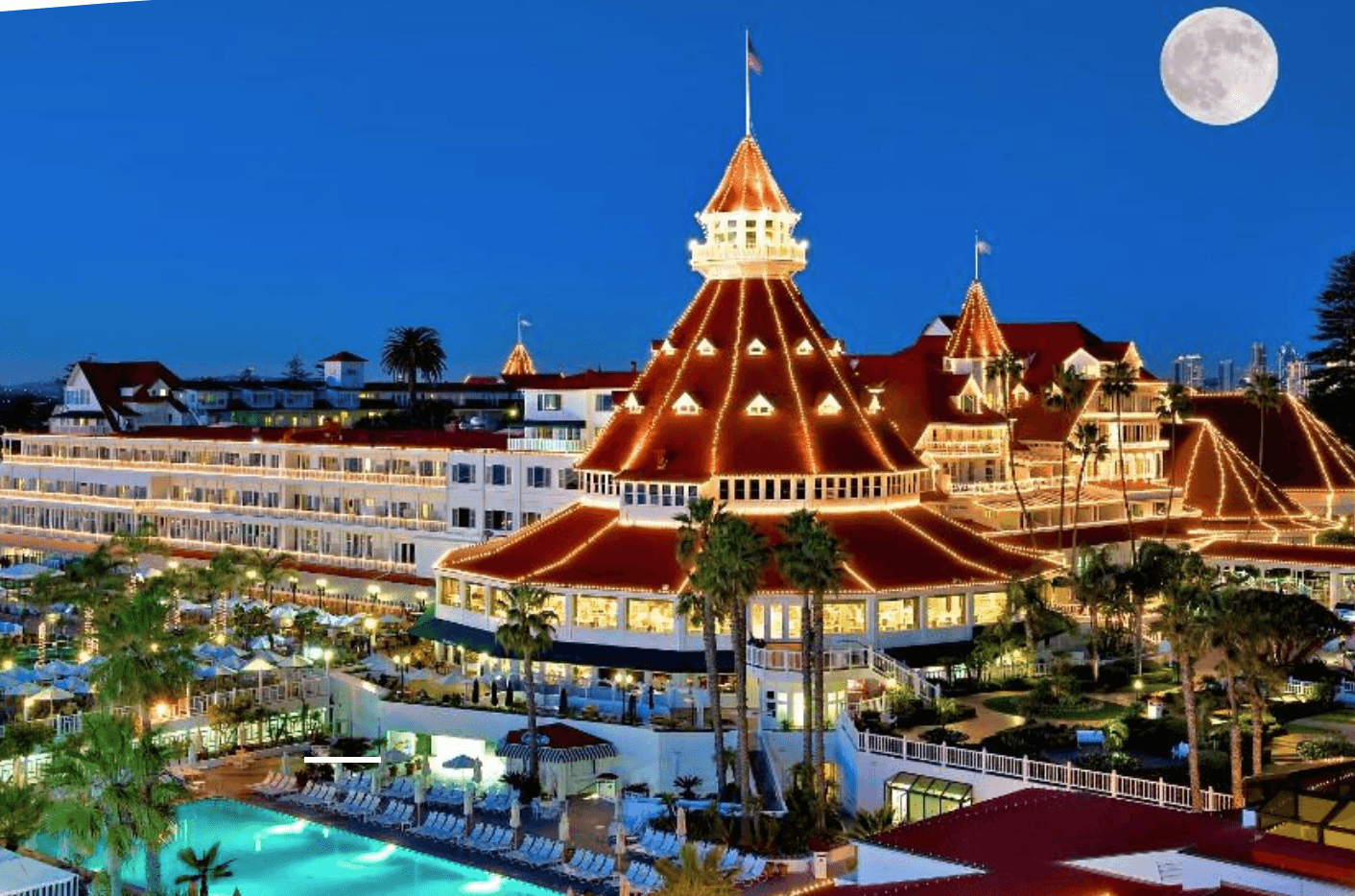 Night view of a large building with illuminated roofs, palm trees, a pool, and a full moon in the sky.
