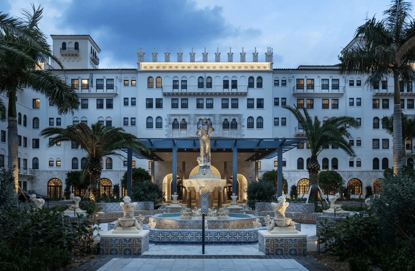 Large white hotel building with palm trees, fountain, and statues in front, under a cloudy sky.