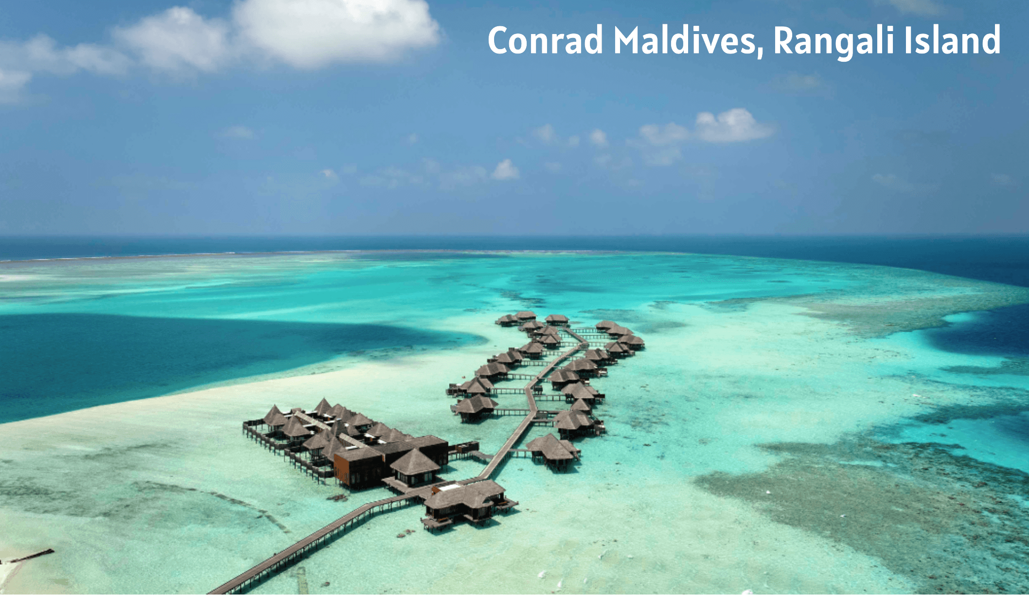 Aerial view of overwater bungalows connected by a walkway in turquoise waters at Conrad Maldives, Rangali Island.