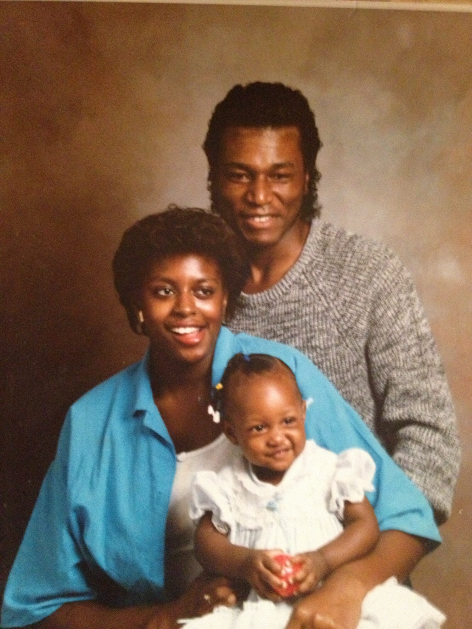 Three people, two adults and a child, smiling and posing together against a brown background.