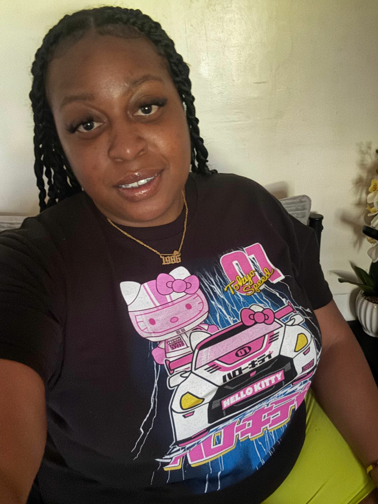 Woman with braided hair smiling, wearing a black T-shirt with colorful car graphic, sitting indoors.