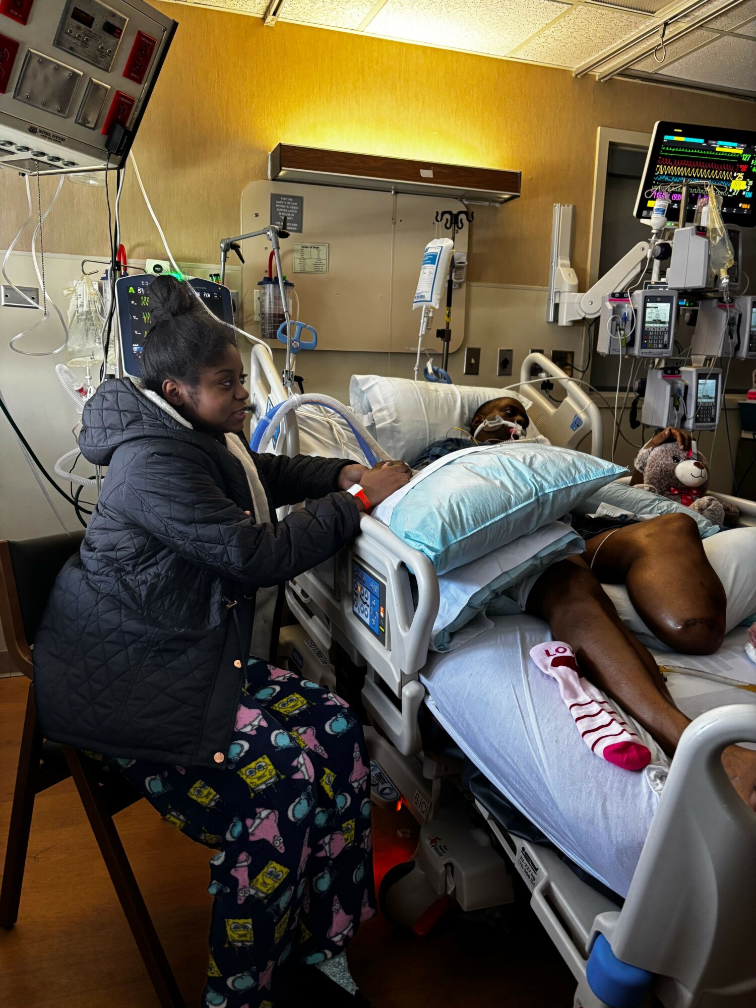 Person lying in hospital bed connected to medical equipment, with a healthcare worker attending, in a hospital room.