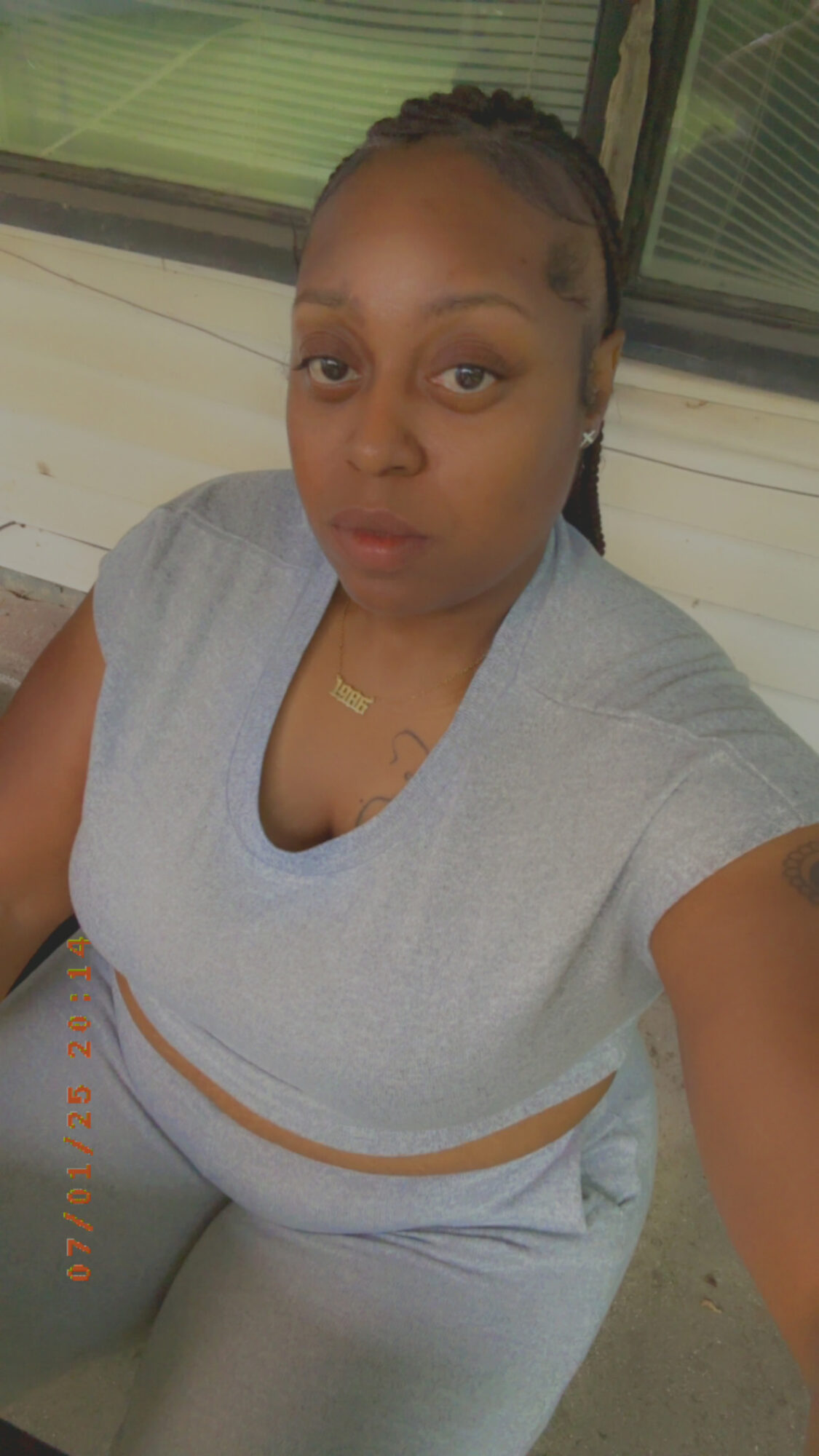 Woman with braided hair sitting outdoors, wearing a light gray top and pants, looking at the camera.