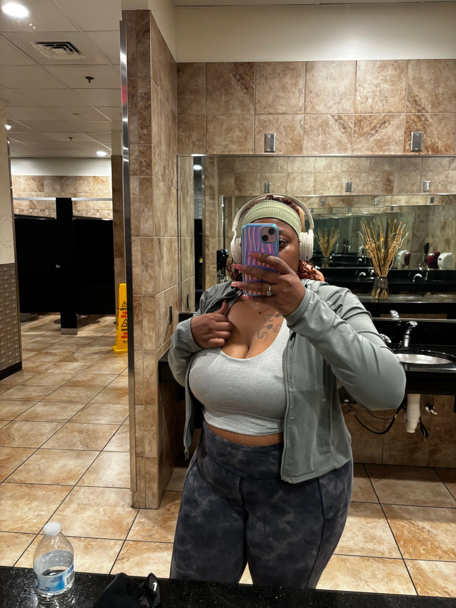 Person taking a mirror selfie in a bathroom, wearing a gray jacket and dark pants, holding a phone with a pink case.