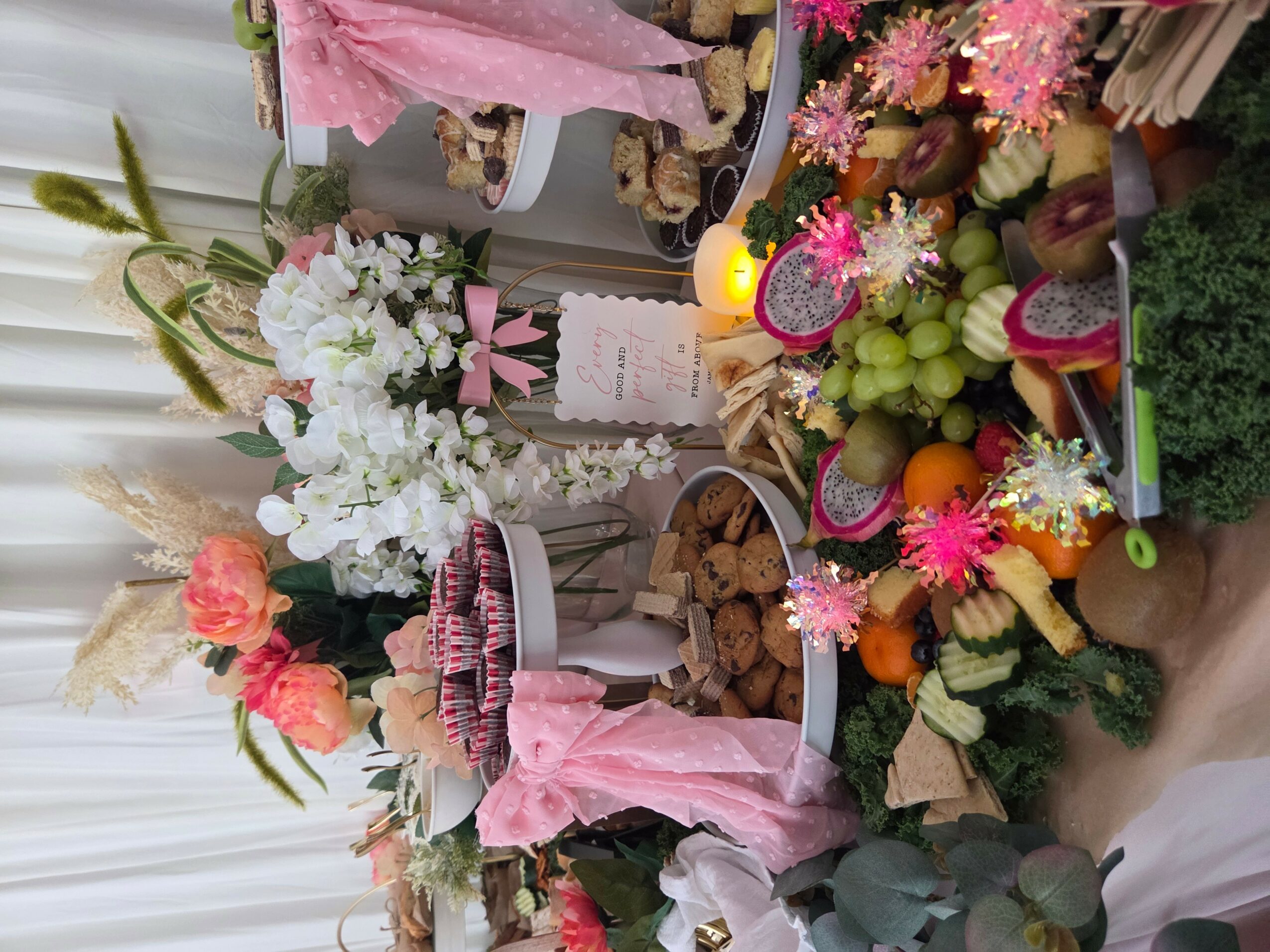 Table with flowers, fruits, desserts, and decorative items, arranged for celebration or event, viewed from the side.