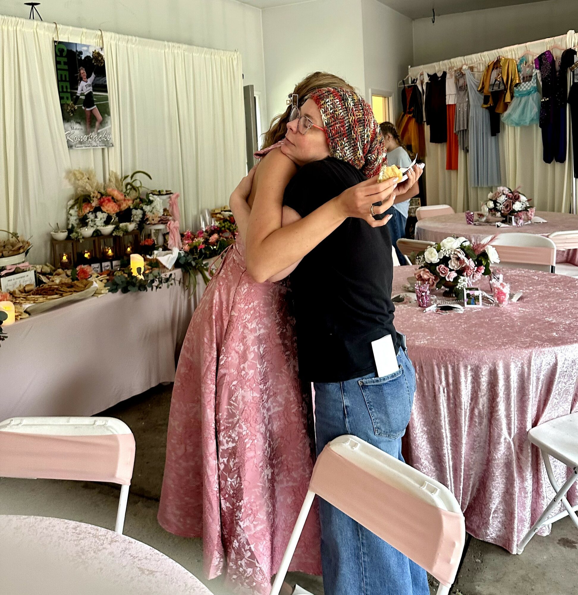Two women hugging in a decorated room with tables, flowers, and clothing racks, one wearing a pink dress and glasses, the other in a black top and jeans.
