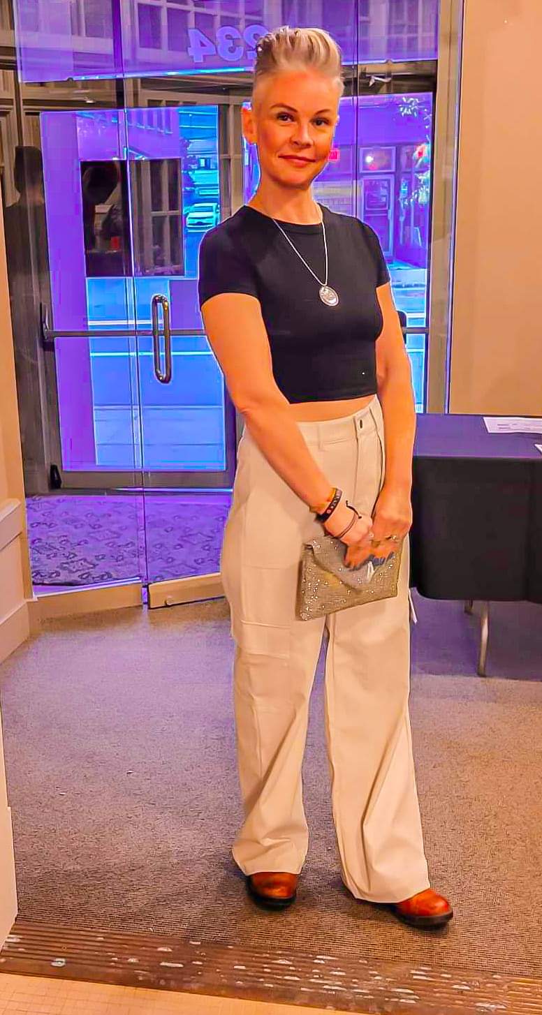 Woman standing indoors, wearing a black top, beige wide-leg pants, holding a small bag, smiling.